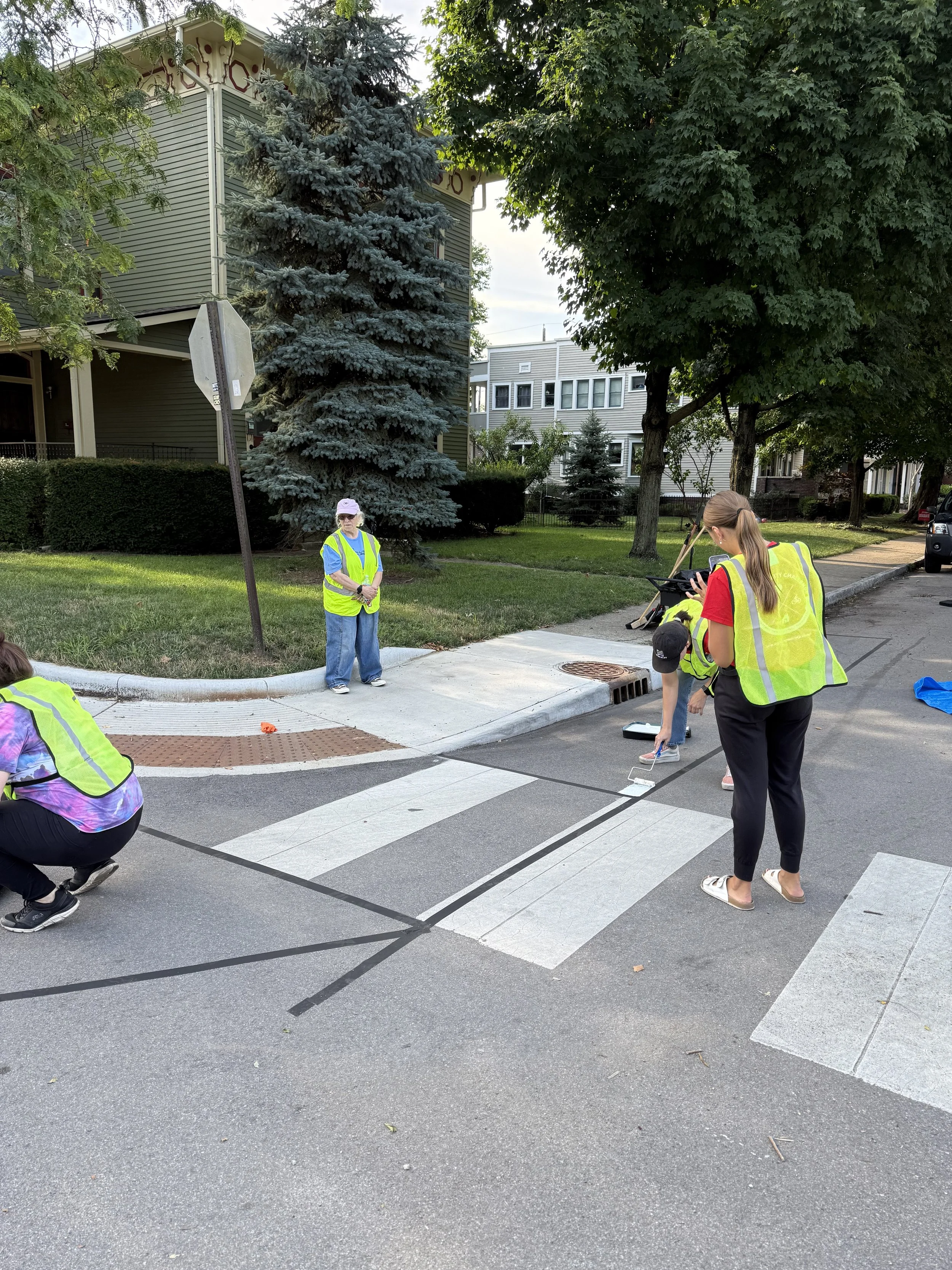 Group of people painting crosswalk lines on a city street, some wearing yellow safety vests, in a residential area with trees and houses in the background.