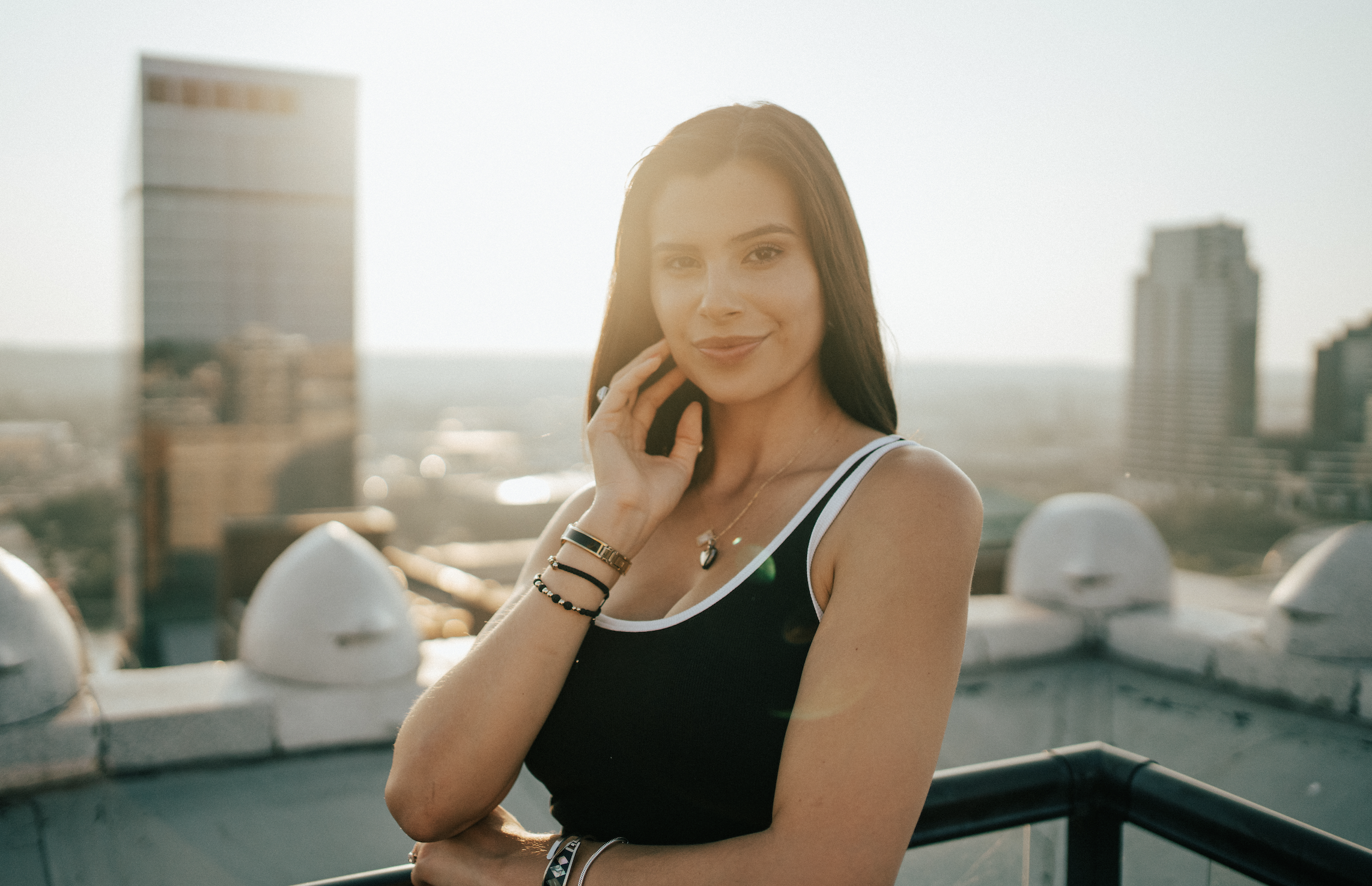 A young woman standing on a rooftop with a city skyline in the background, smiling and touching her face.