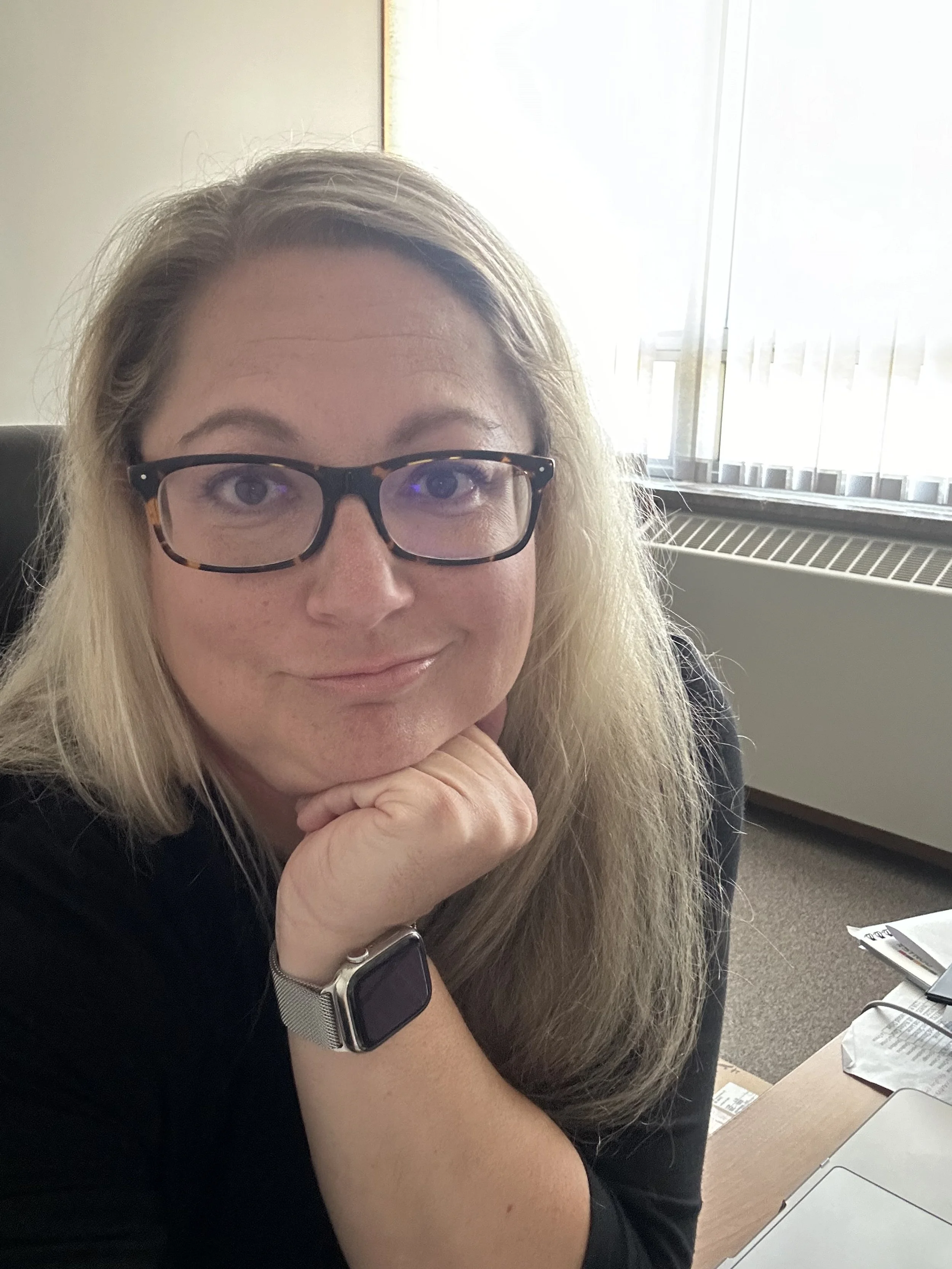 A woman with blonde hair, glasses, and a smartwatch, resting her chin on her hand in an office setting, with a window in the background.