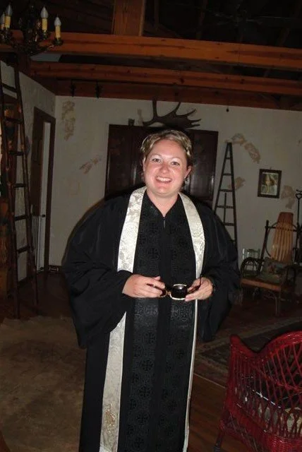 Person in graduation gown and stole standing indoors, smiling, with rustic decor including a ladder and rocking chair in the background.