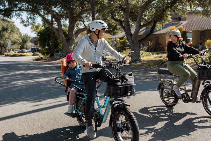 A family man riding an electric bicycle with two children, one in a child seat and the other on a separate bike, in a suburban neighborhood near Rochester NY with trees and houses.