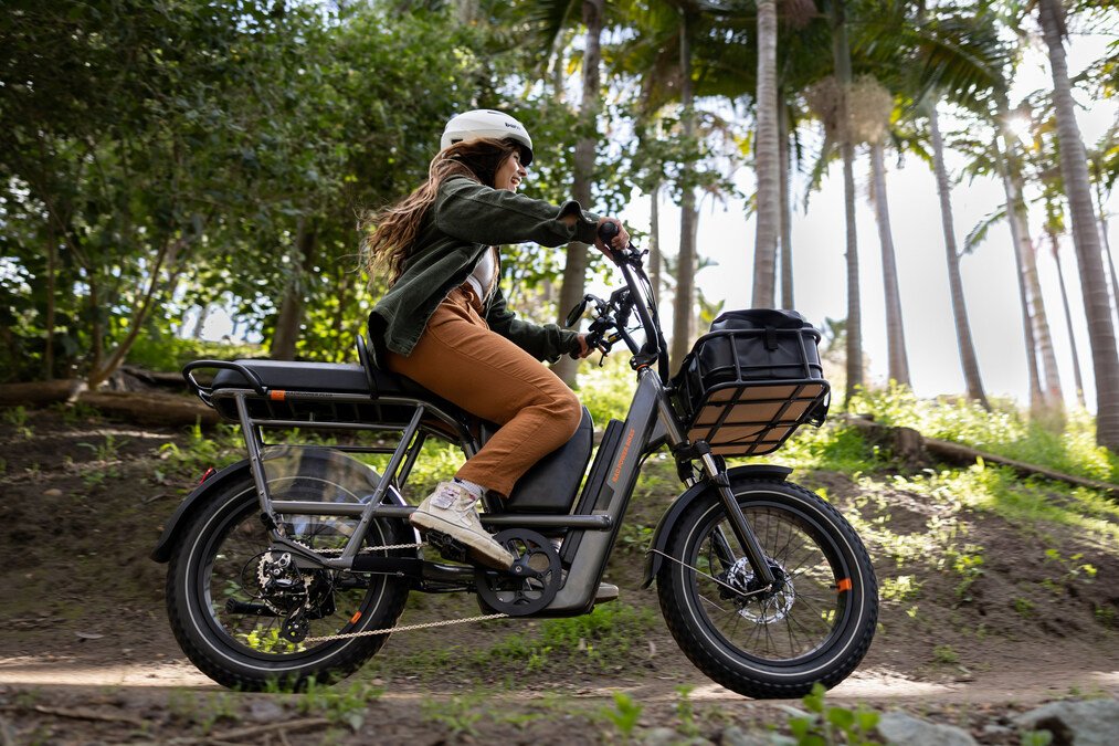 A woman wearing a helmet riding a black electric bike on a forest trail near Pittsford NY surrounded by tall trees and sunlight.