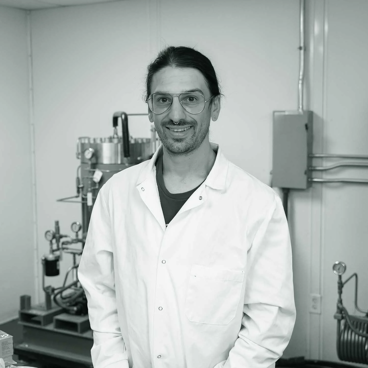 A male scientist in a white lab coat standing in a laboratory, smiling at the camera.