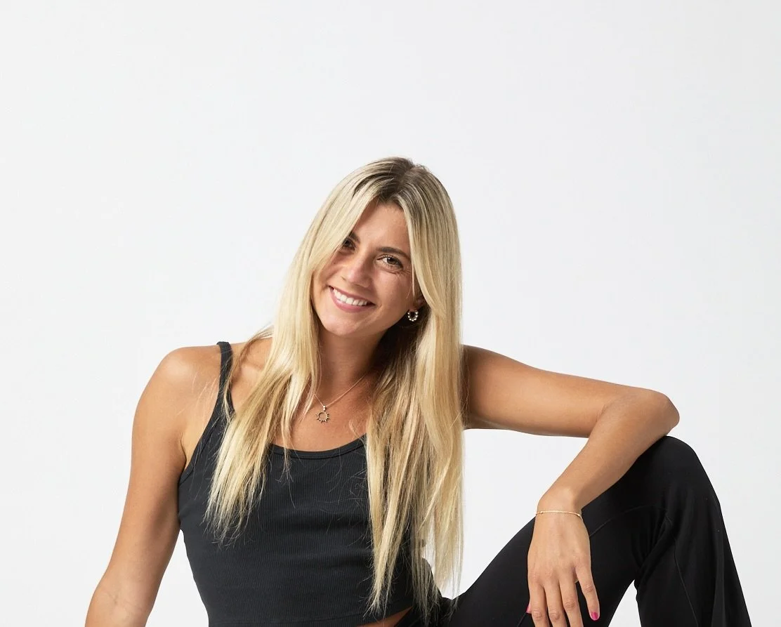 A young woman with long blonde hair, smiling, wearing a black tank top and black pants, sitting against a white background.