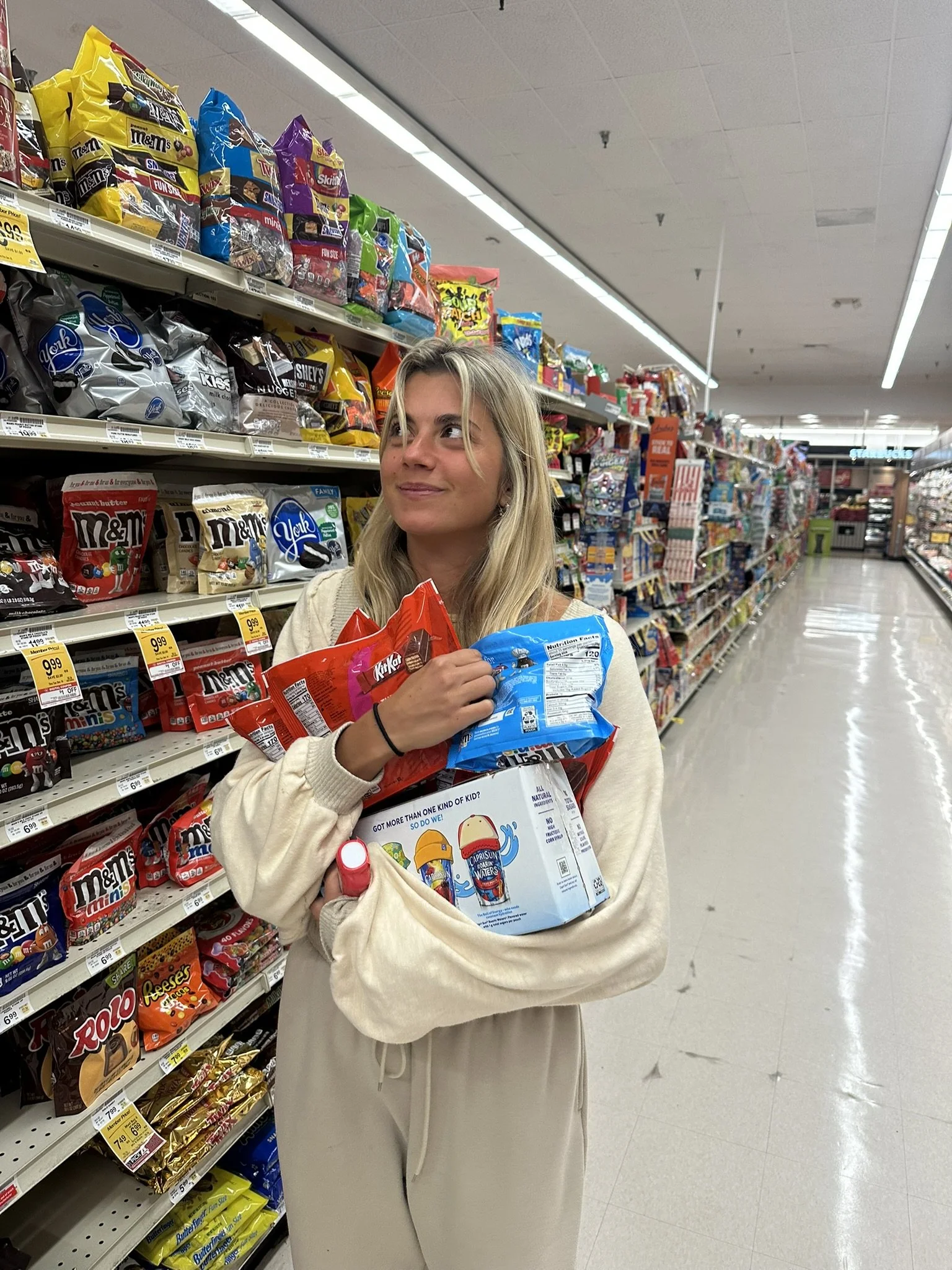 A young woman standing in an aisle of a grocery store, holding multiple packs of snack candies and a palette with a character on it, looking to the side with a slight smile.