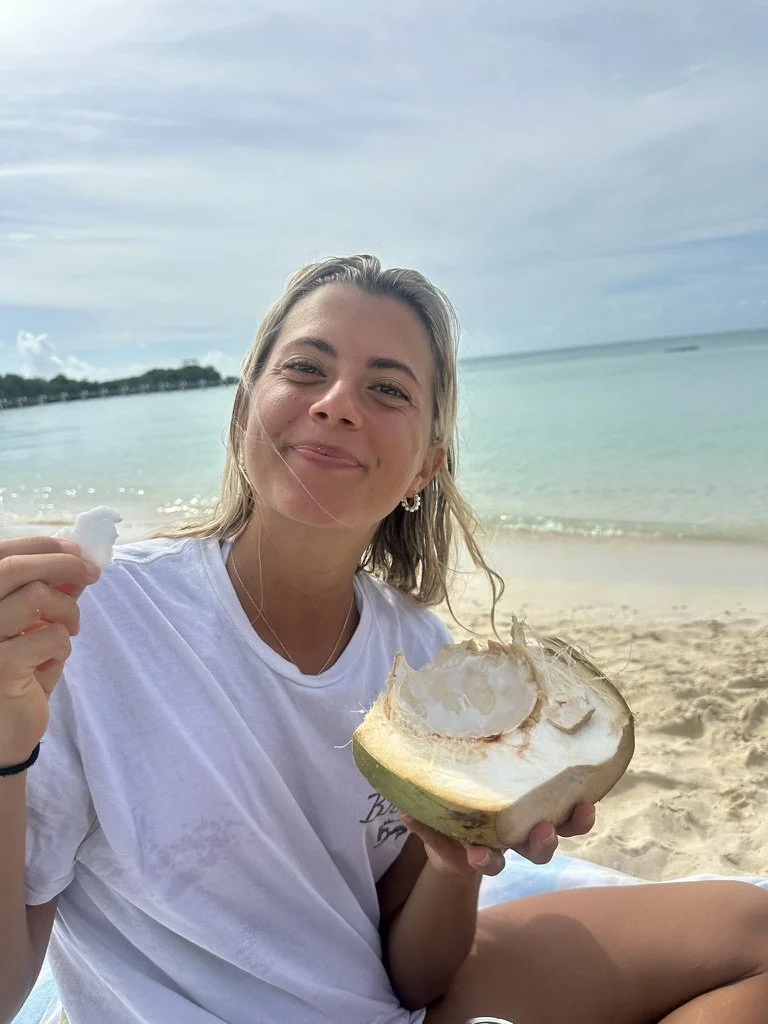 A smiling woman sitting on a beach holding a half-eaten coconut in one hand and a piece of coconut meat in the other, with the ocean and cloudy sky in the background.