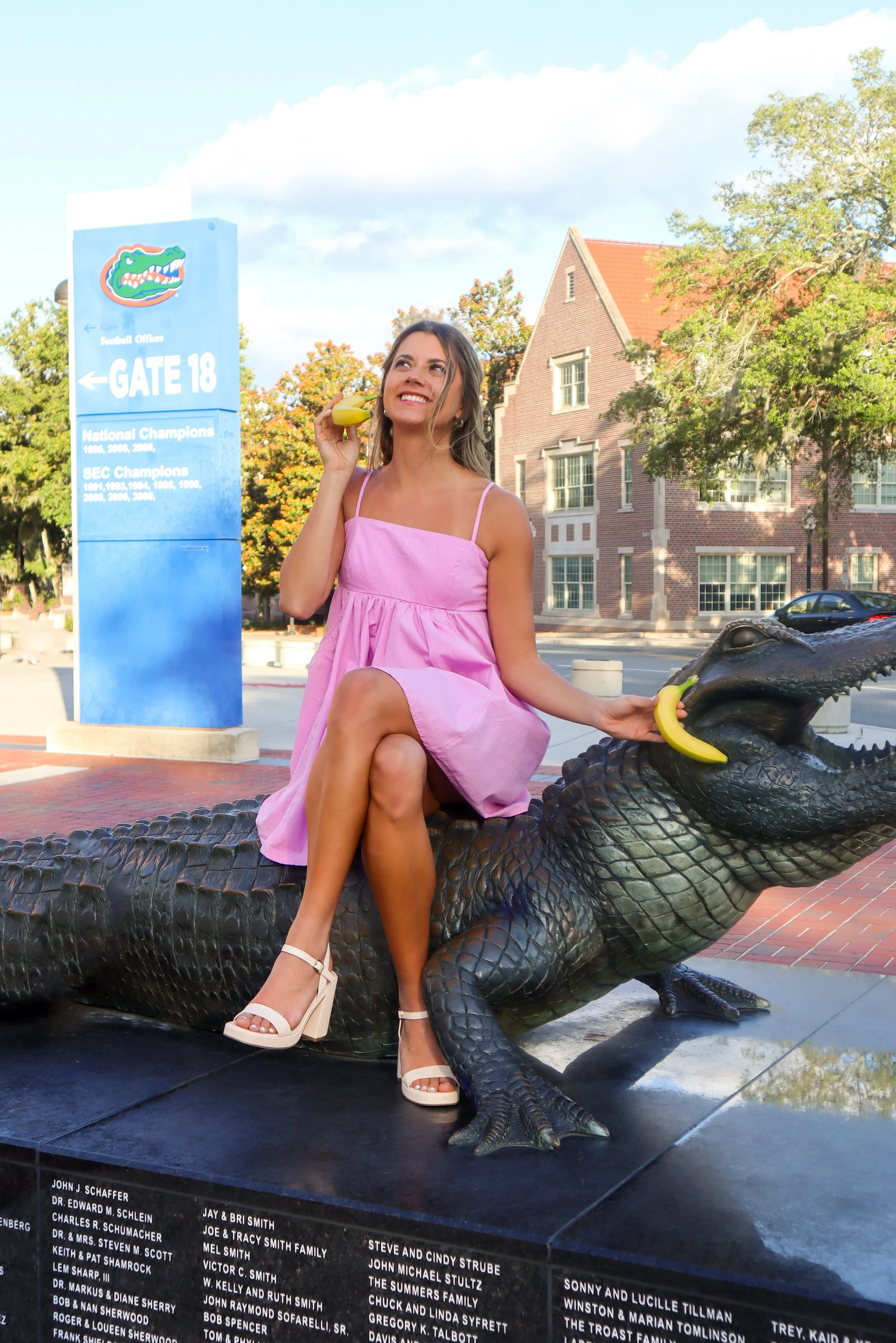 A woman in a pink dress sitting on an alligator sculpture, holding a banana to her ear like a phone, in front of a sign showing the Gator logo and a building.