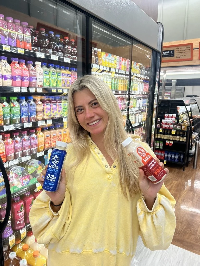A woman with blonde hair wearing a yellow sweatshirt shopping for probiotics in a grocery store, holding a blue probiotic bottle in her right hand and a red probiotic bottle in her left hand, in front of refrigerated shelves with various flavored drinks.