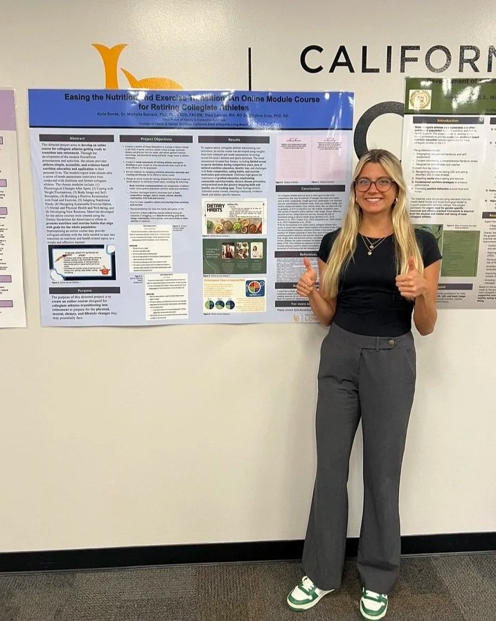 Young woman with long blonde hair, glasses, black t-shirt, gray pants, and green sneakers giving a thumbs-up while standing next to an academic research poster on a conference wall.