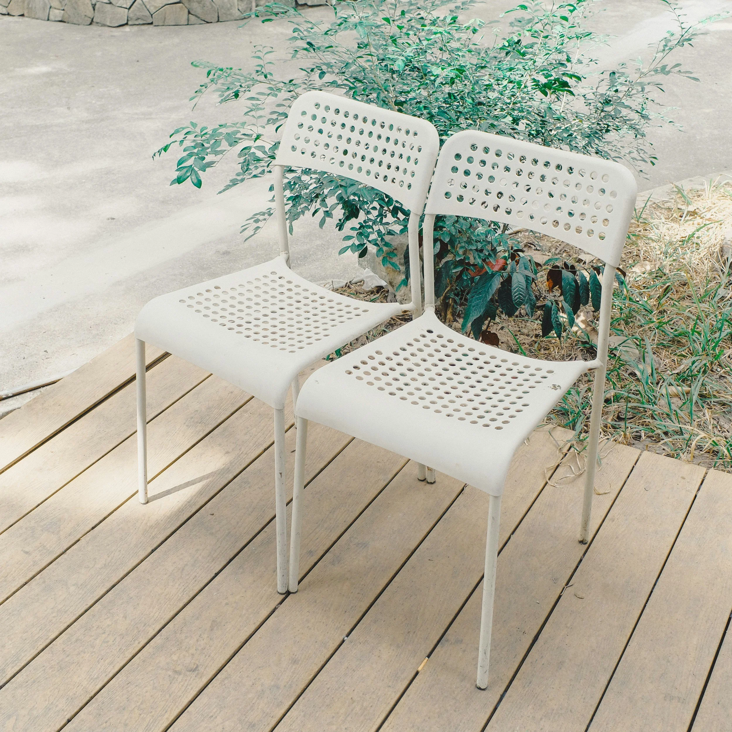 Two white patio chairs set up beside one another with a bush in background.