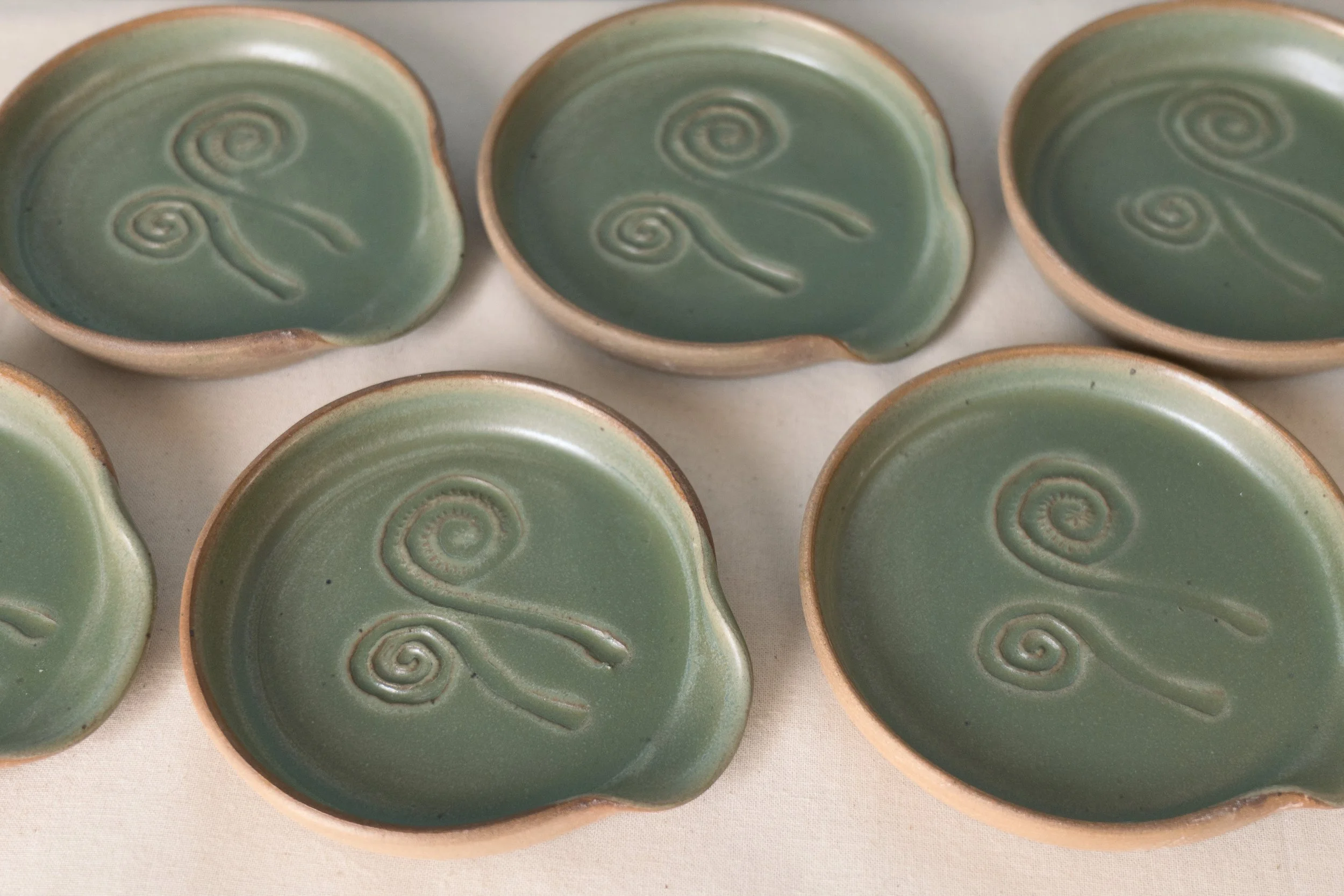 Seven small, round, green ceramic spoon rests with fiddlehead designs inside, arranged on a light-colored surface. Copyright Kate O'Rourke/Maritime Ceramics.
