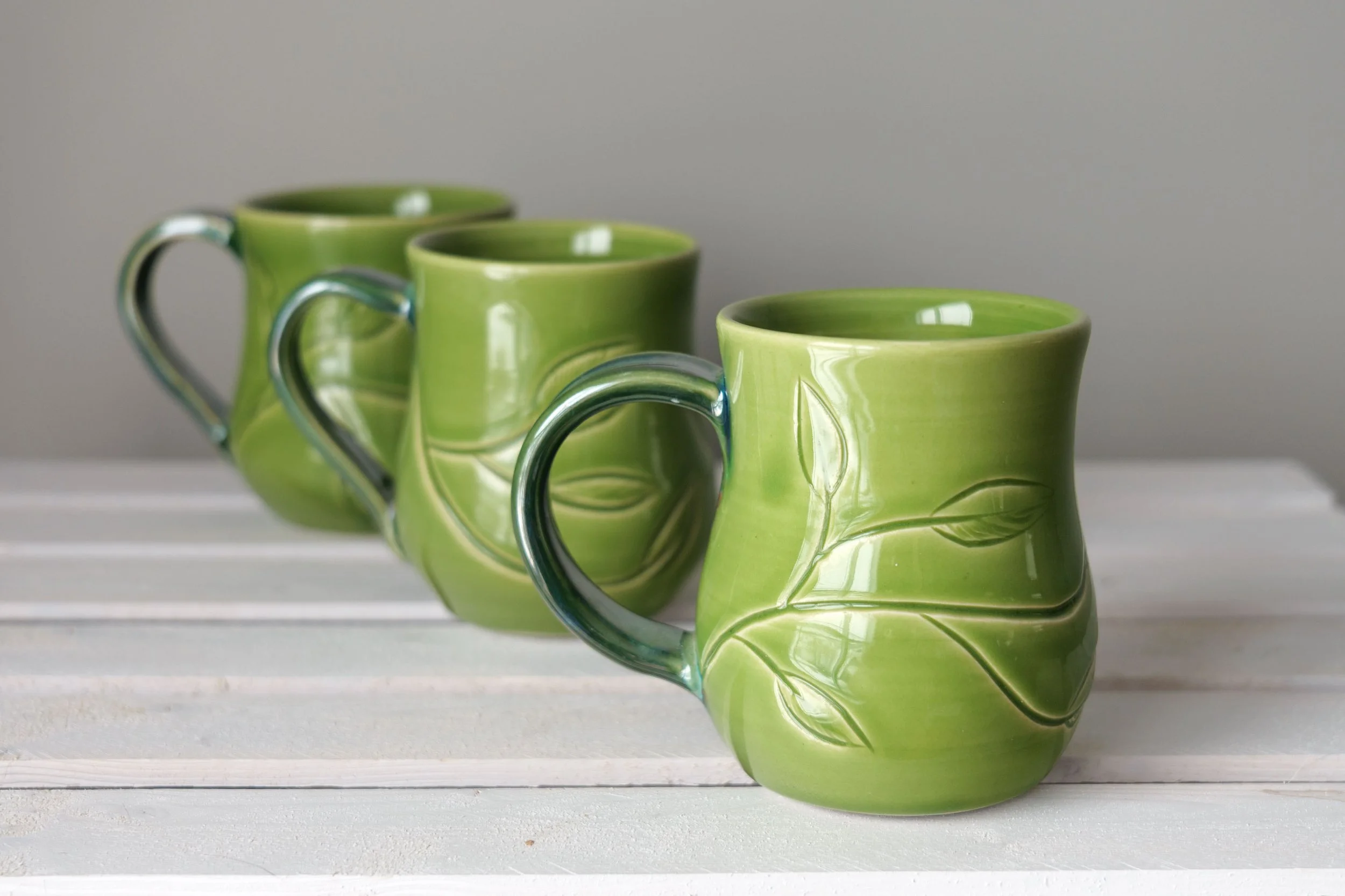 Three green mugs with leafy vines carved into the surface on a white wood surface with a grey background