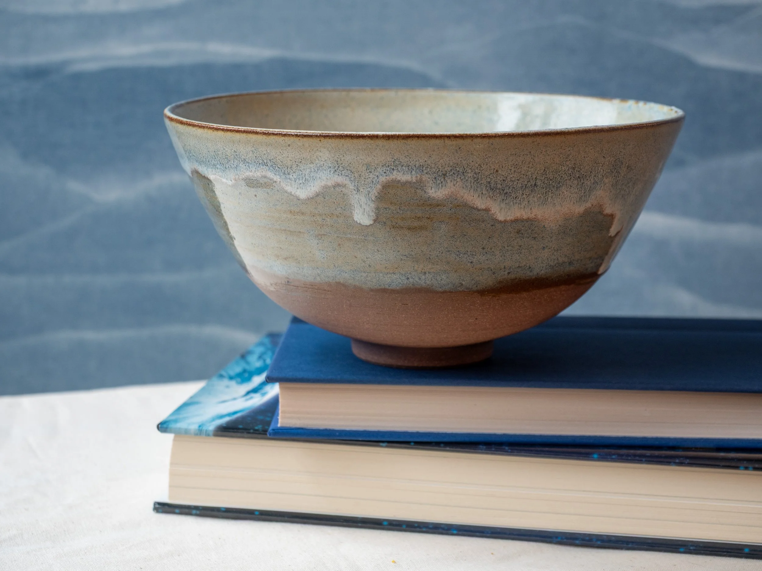 A ceramic bowl with a textured glaze sits on top of two stacked books with a blue cover. The background consists of a light blue and white wavy pattern.