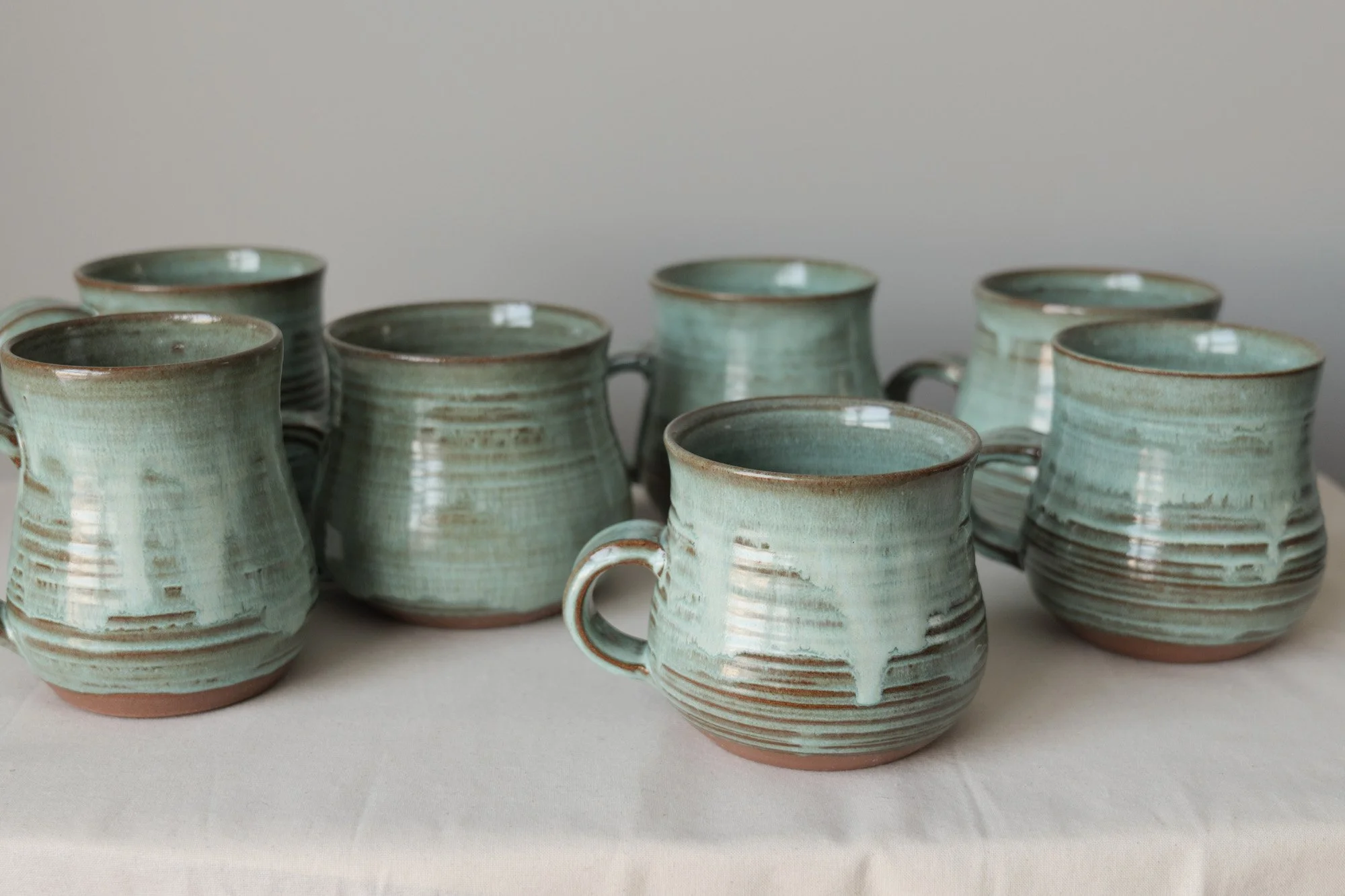 Seven handmade ceramic coffee mugs with a glossy green glaze and horizontal ribbed texture, arranged on a white fabric surface against a plain background. Copyright Kate O'Rourke/Maritime Ceramics.