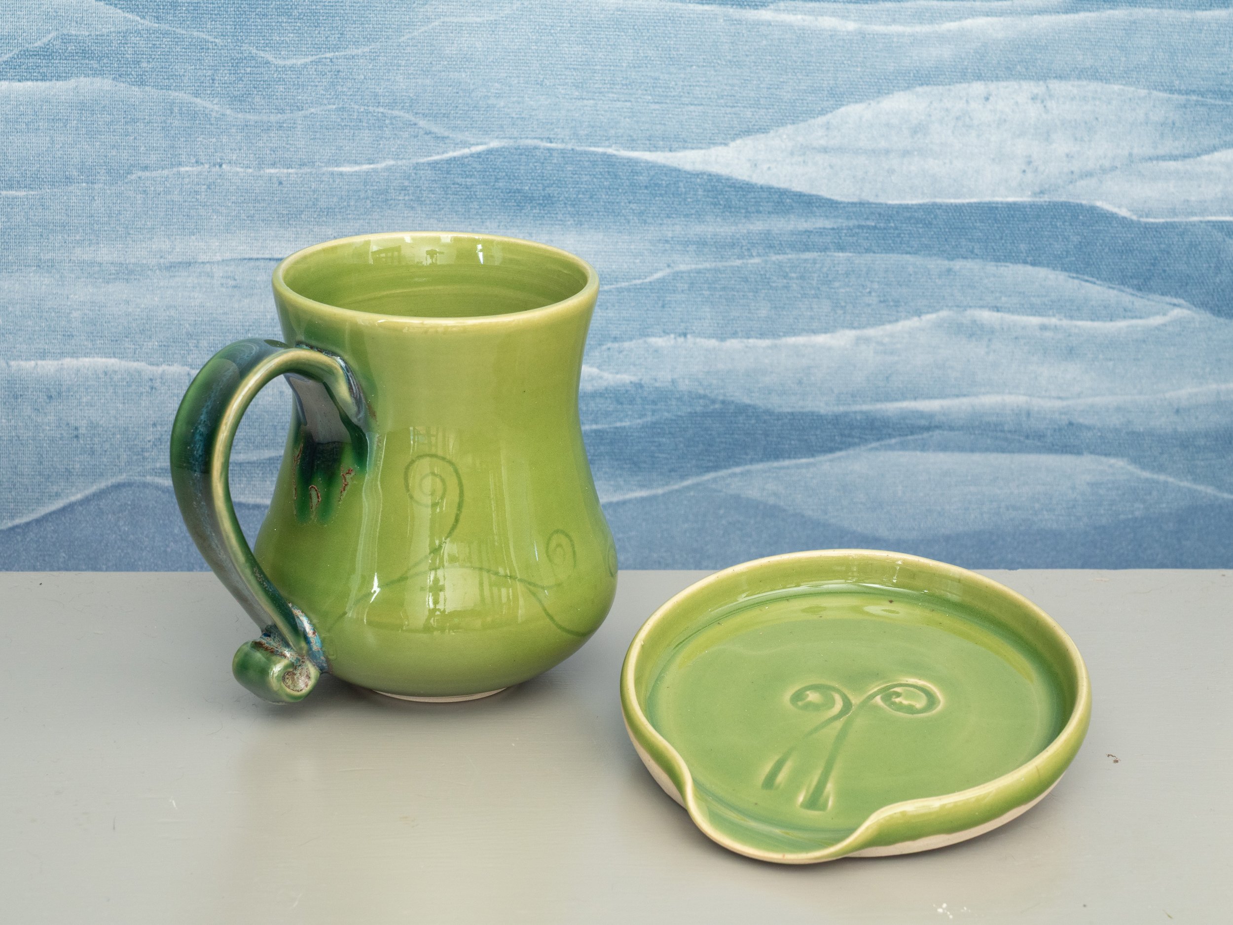 A green ceramic mug and matching spoon rest with swirling vine designs on a light gray surface, against a blue textured background.