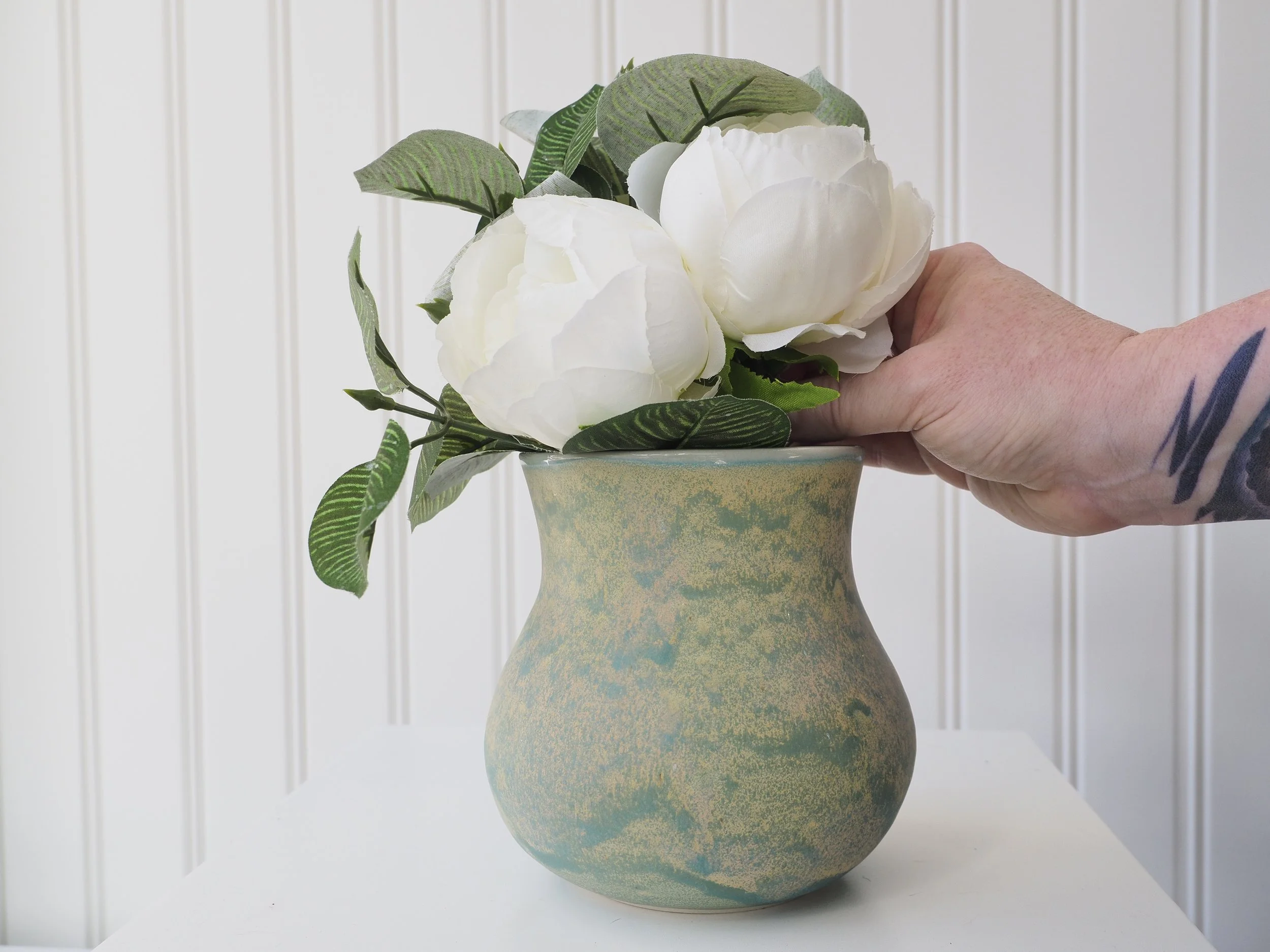 Hand arranging white peony flowers with green leaves in a textured, two-toned yellow and blue ceramic vase, against a white paneled wall background.
