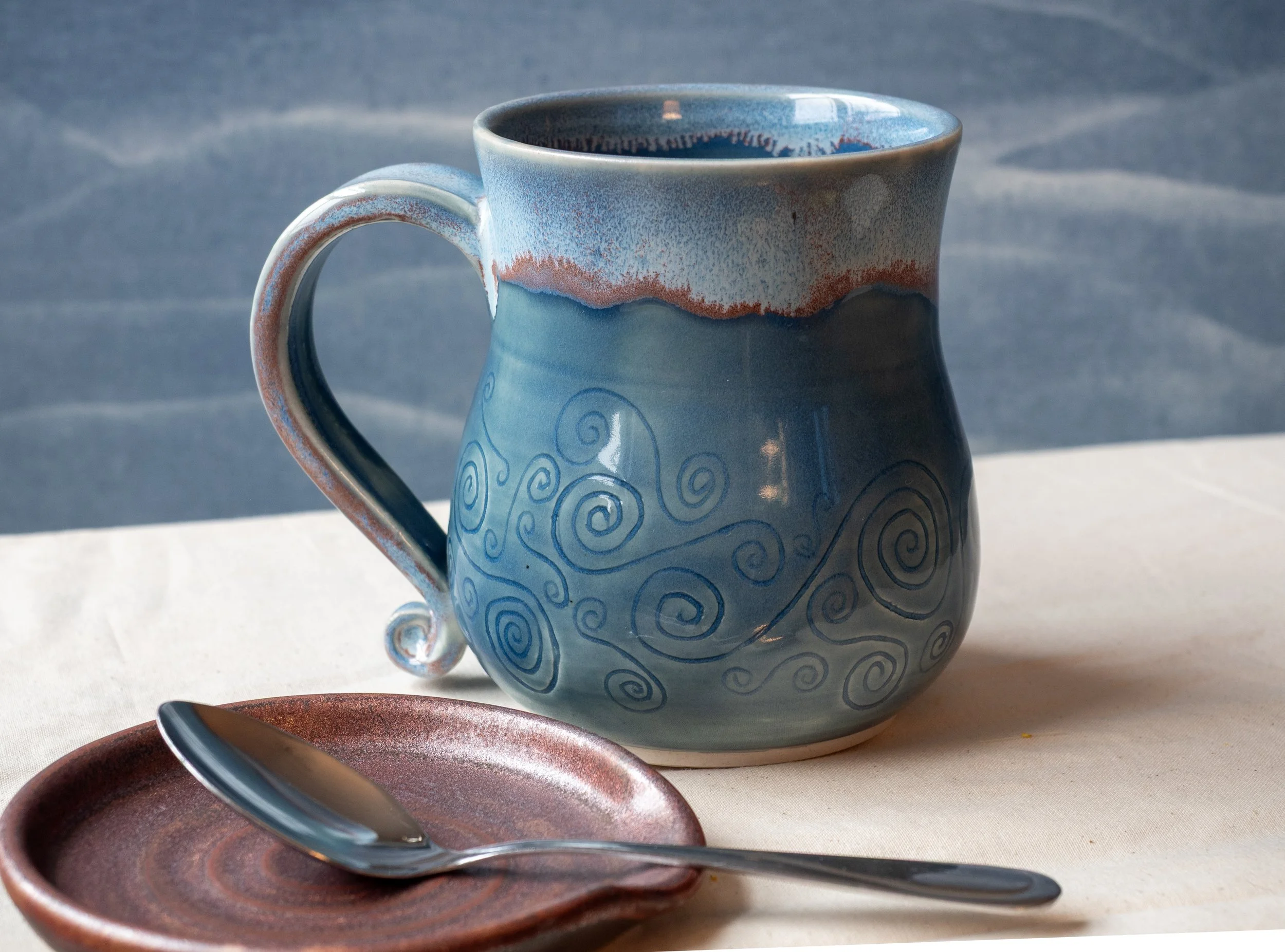 A ceramic mug with a blue and white glaze, decorated with spiral patterns, placed on a table with a small copper spoon rest and a silver spoon.