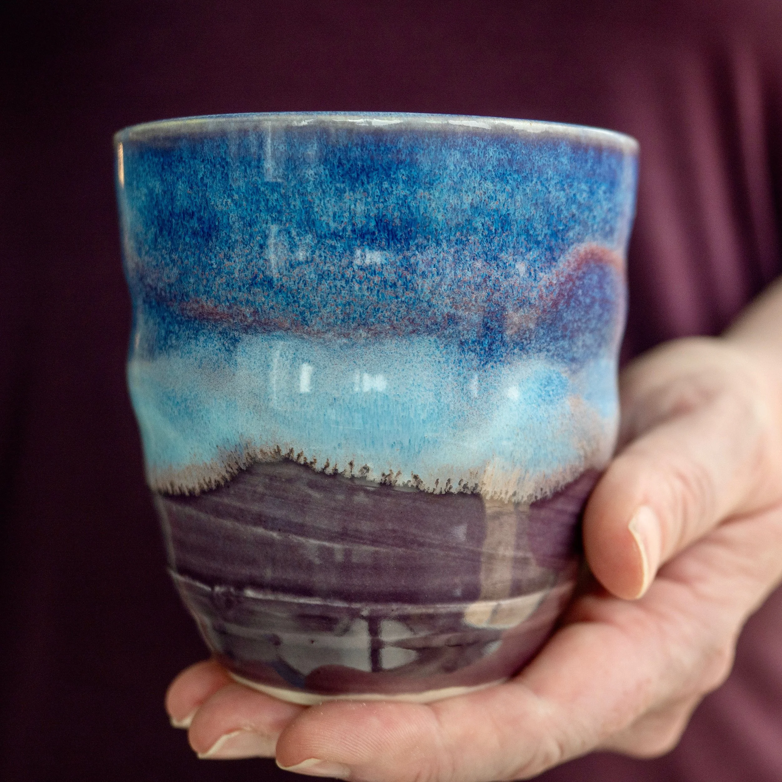 A hand holding a ceramic cup with a blue and purple glaze pattern resembling a landscape with blue sky, clouds, and distant mountains. Copyright Kate O'Rourke/Maritime Ceramics.