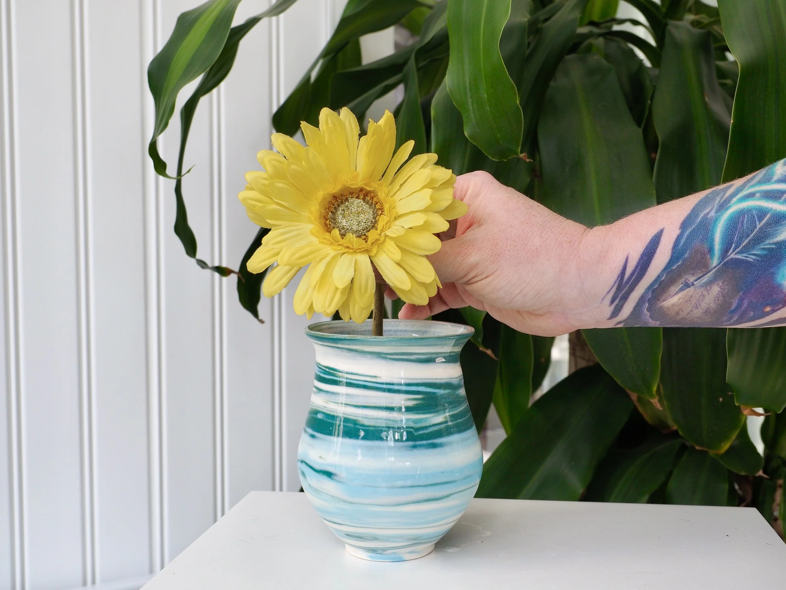 A person with a colorful tattooed arm placing a yellow flower in a blue and white neriage ceramic vase on a white surface, with large green leafy plants in the background.