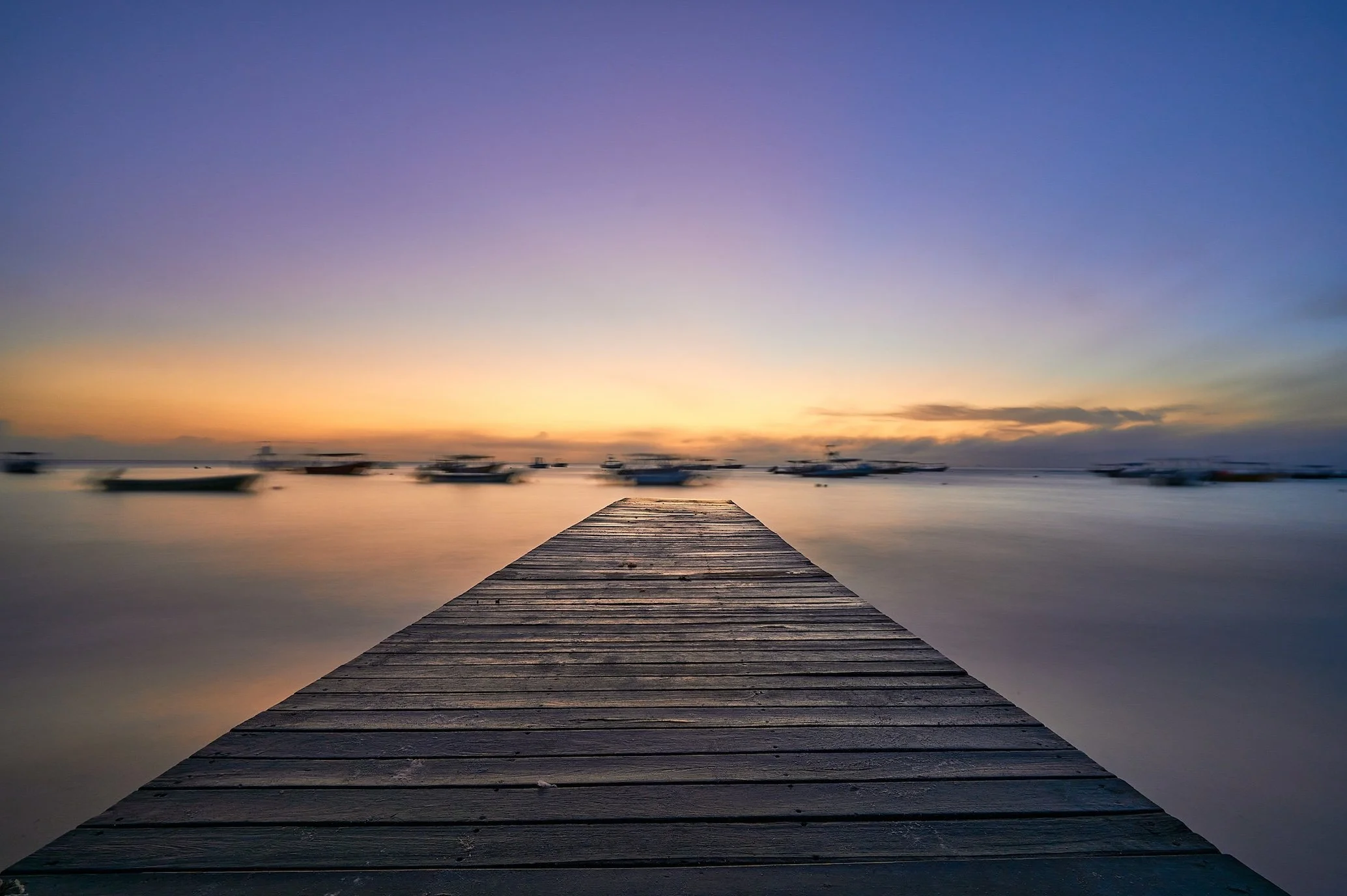 A wooden dock extending into calm water at sunset with boats on the horizon and a colorful sky.
