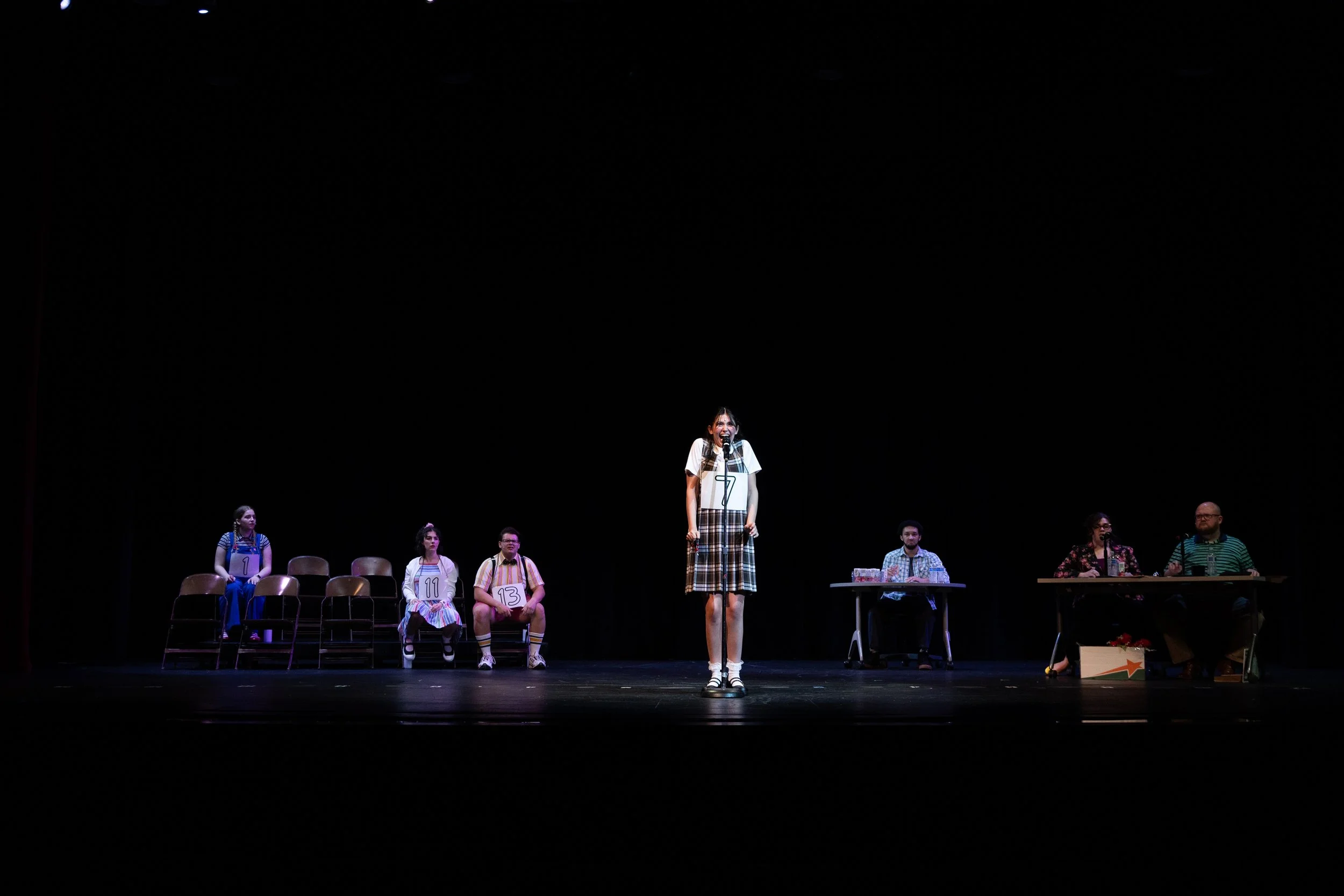 A group of people on a stage, with one woman standing and speaking into a microphone, wearing a plaid skirt and white top. The others sit in chairs or stand at a table, wearing colored bibs with numbers, in a dark theater setting.