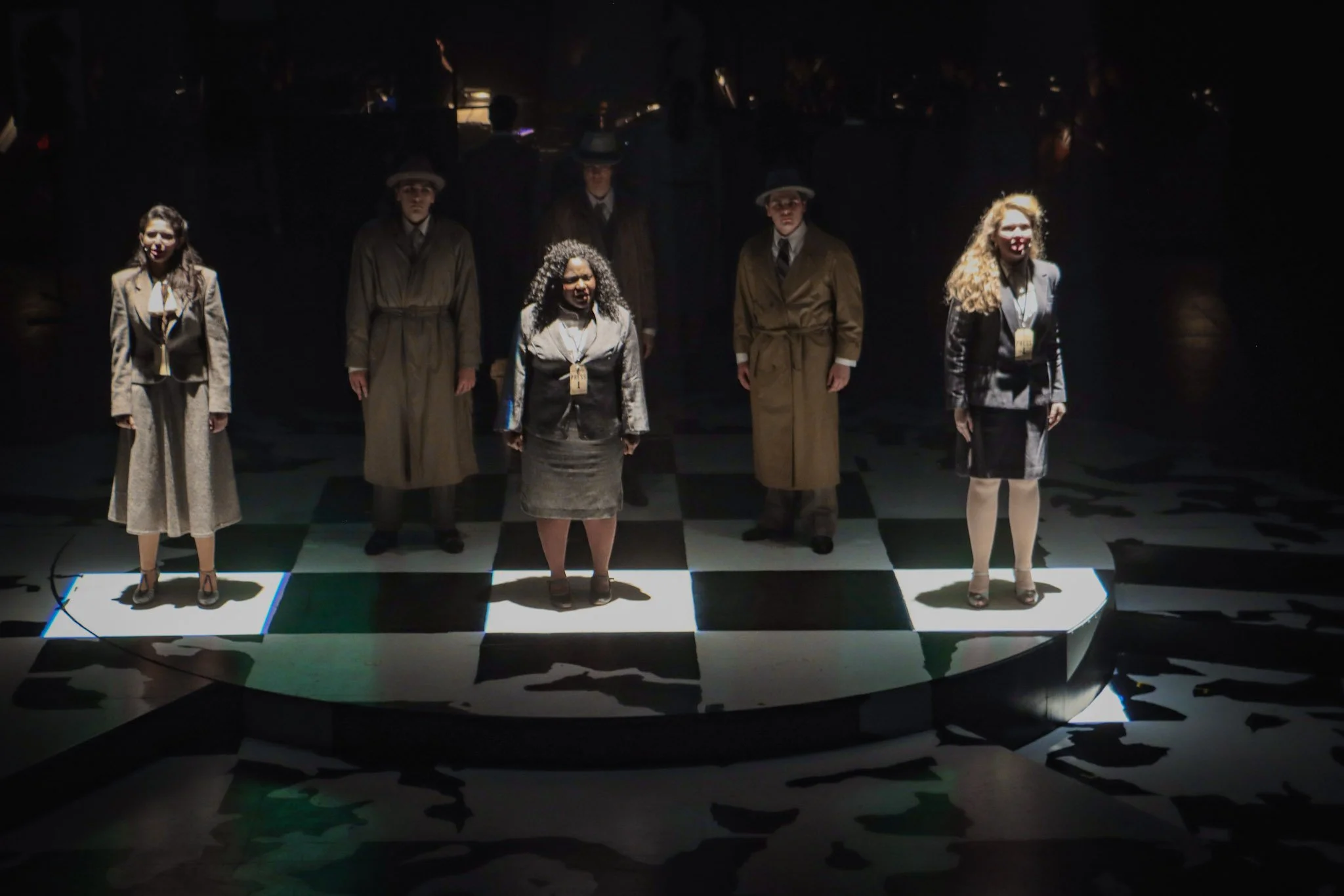A group of seven people standing on a chessboard-like platform in a dark room with dramatic lighting. They are dressed in vintage or formal attire, with some wearing coats and hats, and appear to be part of a theatrical or performance setting.