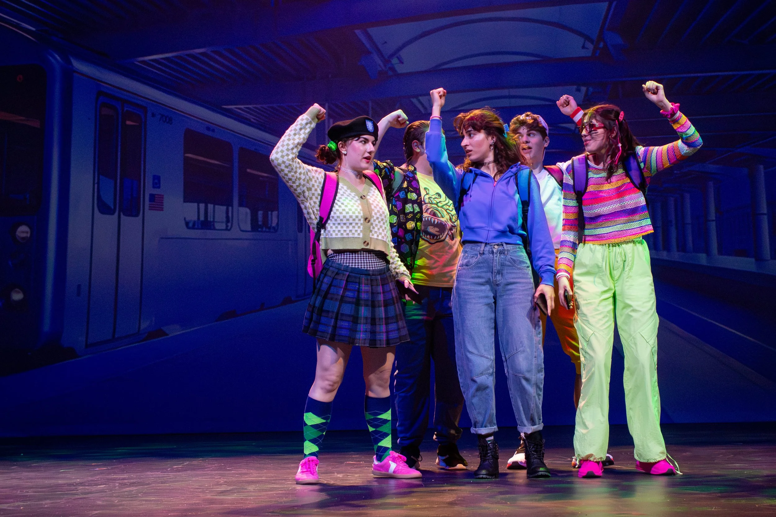 Group of five young women standing on a stage, wearing colorful outfits and backpacks, with a blue train in the background.