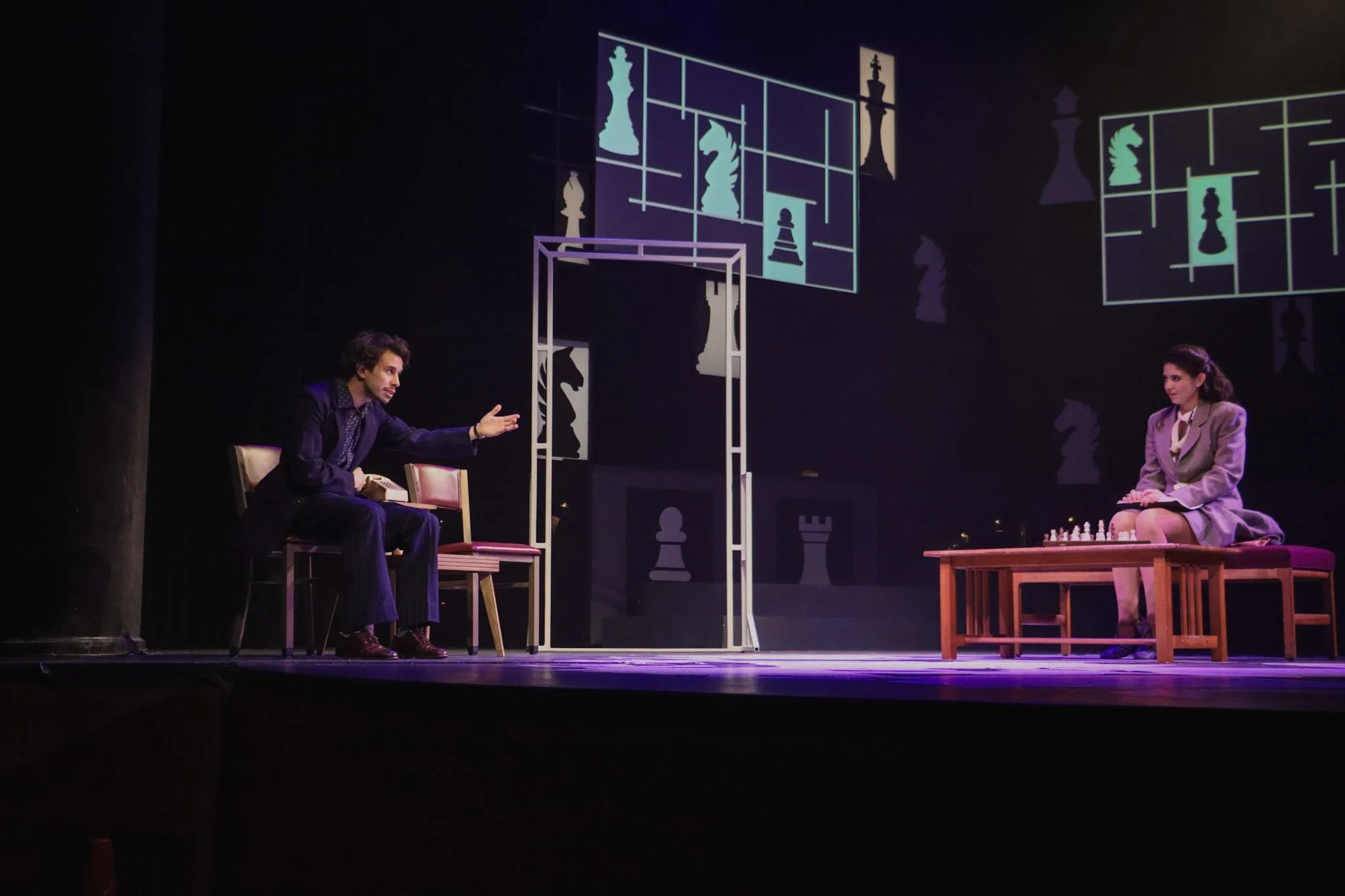 Two actors on stage engaged in a chess-themed theatrical performance, with large chess piece silhouettes projected on the background.