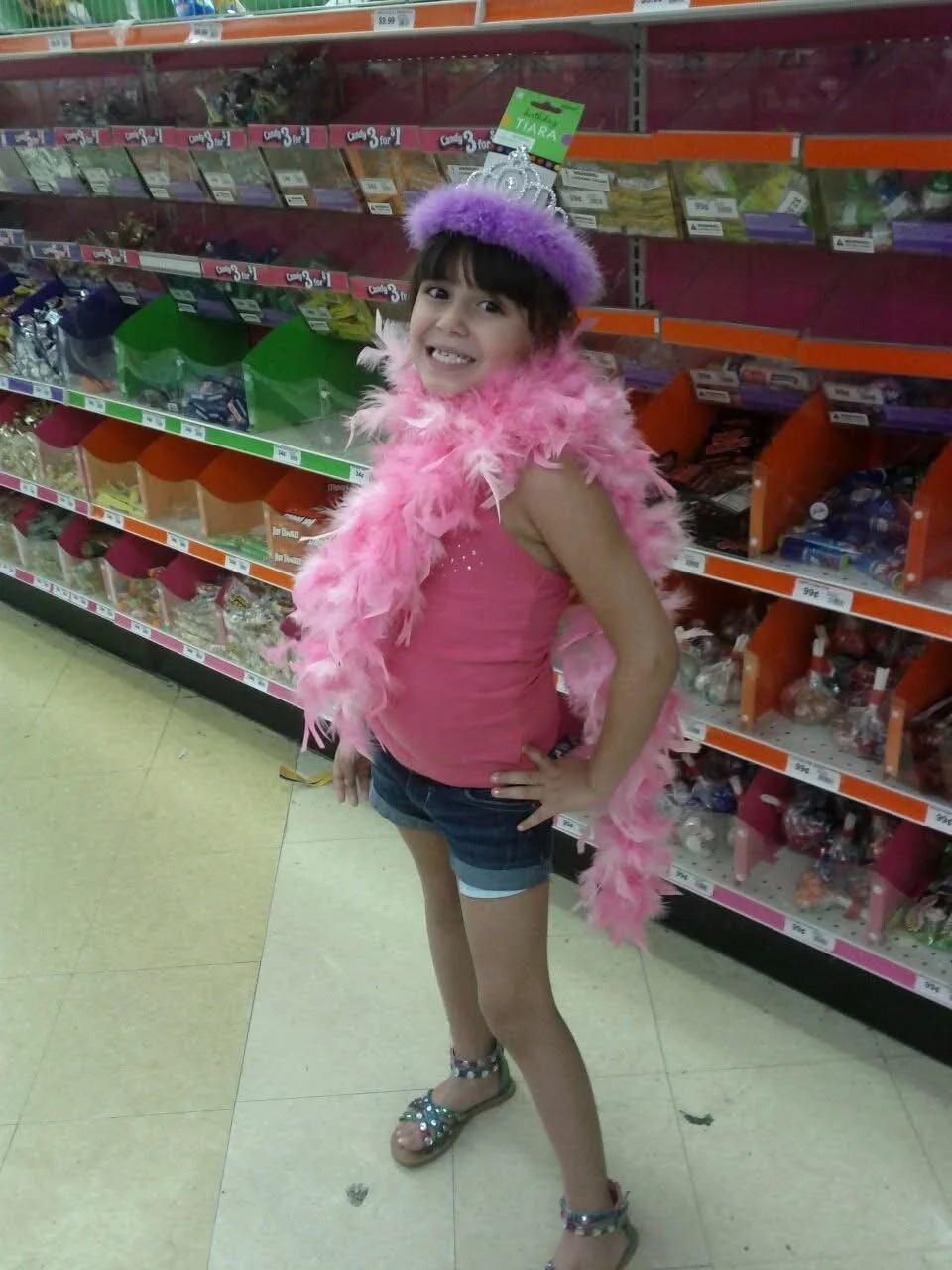 A girl wearing a pink top, denim shorts, and sandals with rhinestones is standing in a store aisle. She is smiling, with her hands on her hips, wearing a purple feathered crown and a pink feather boa, and a tiara on her head.