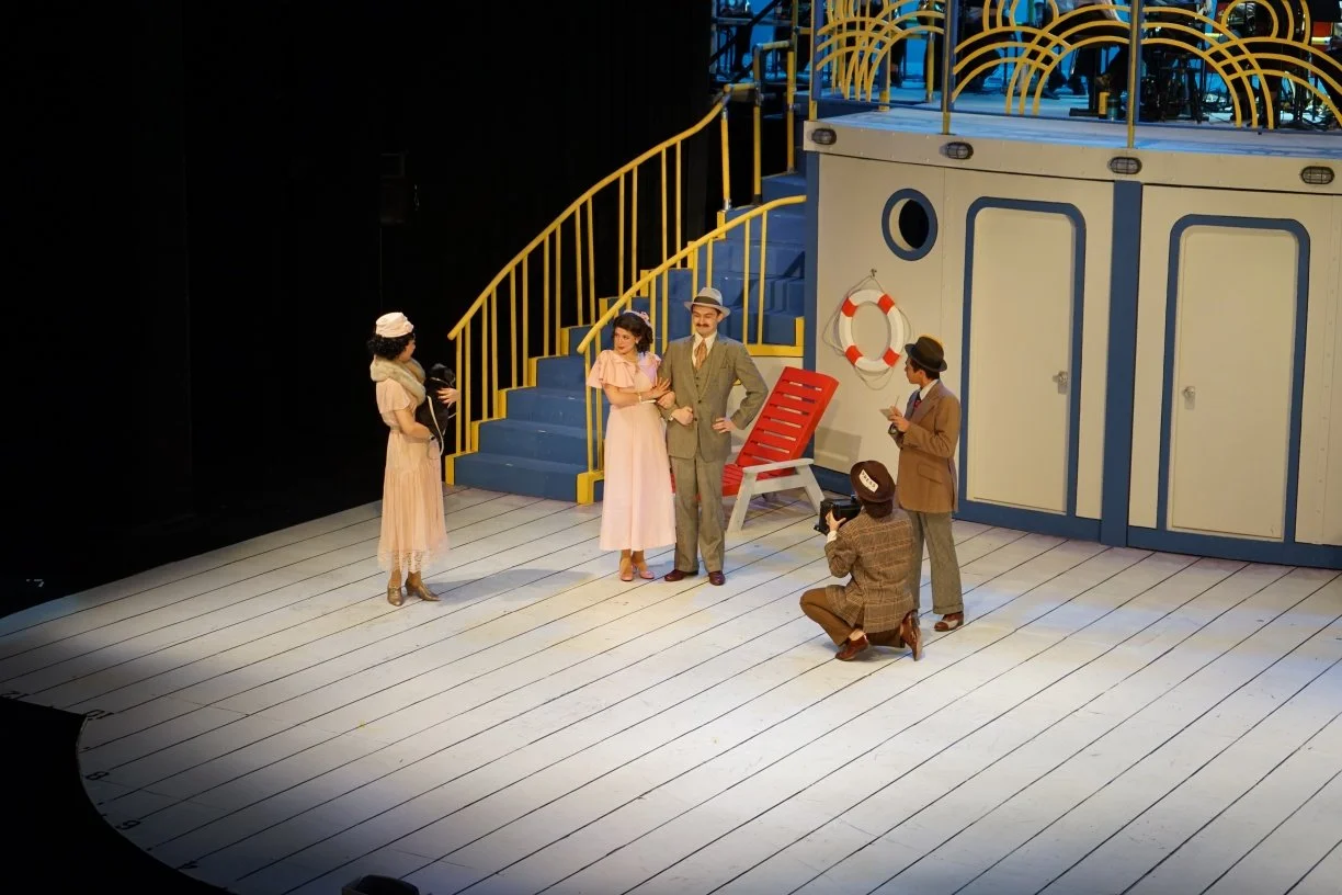 People on a stage set designed to look like a ship deck, with five actors, two of whom are being photographed, one filming, and one standing and talking, all dressed in vintage clothing from the early 20th century.