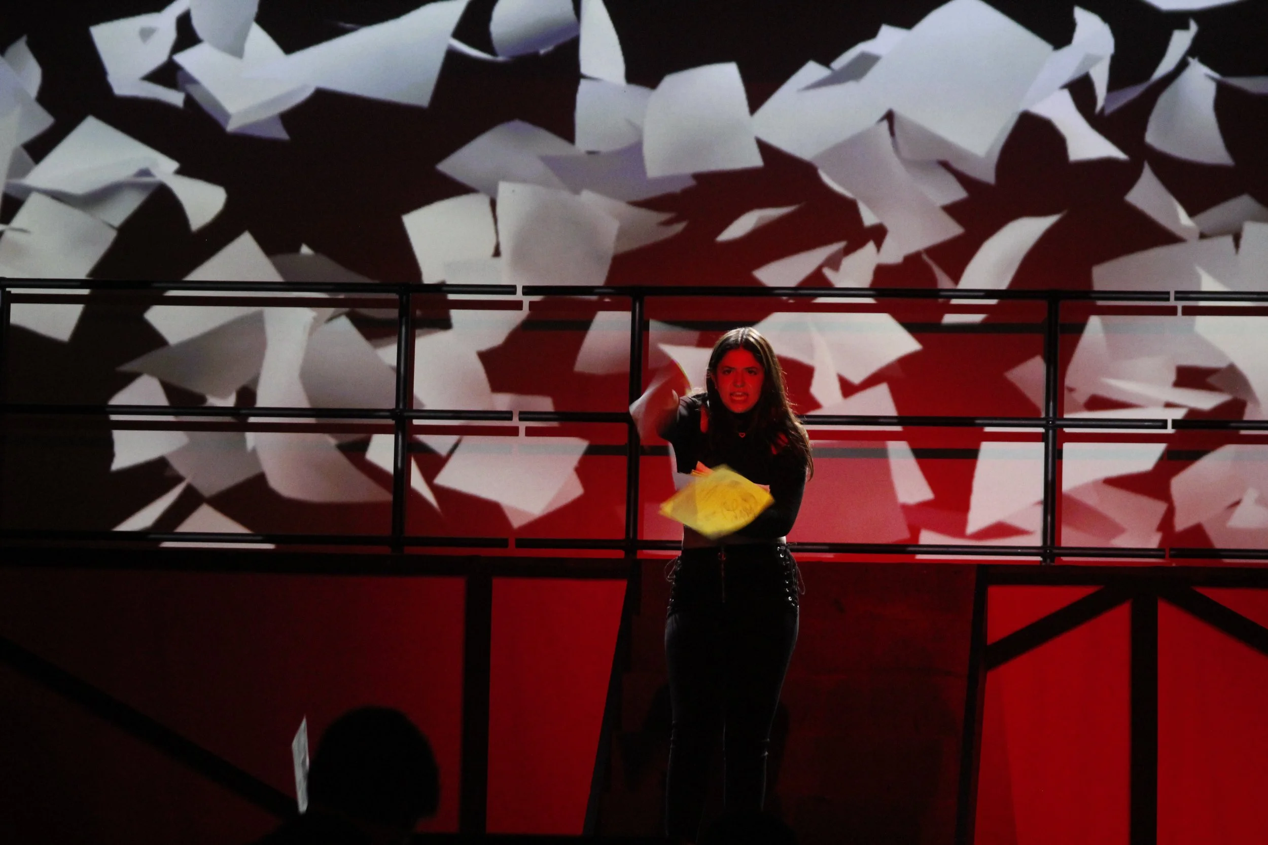 A woman standing on a stage with a background of abstract paper cutouts, holding a yellow paper in her hand, with a red lighting that adds a dramatic effect.