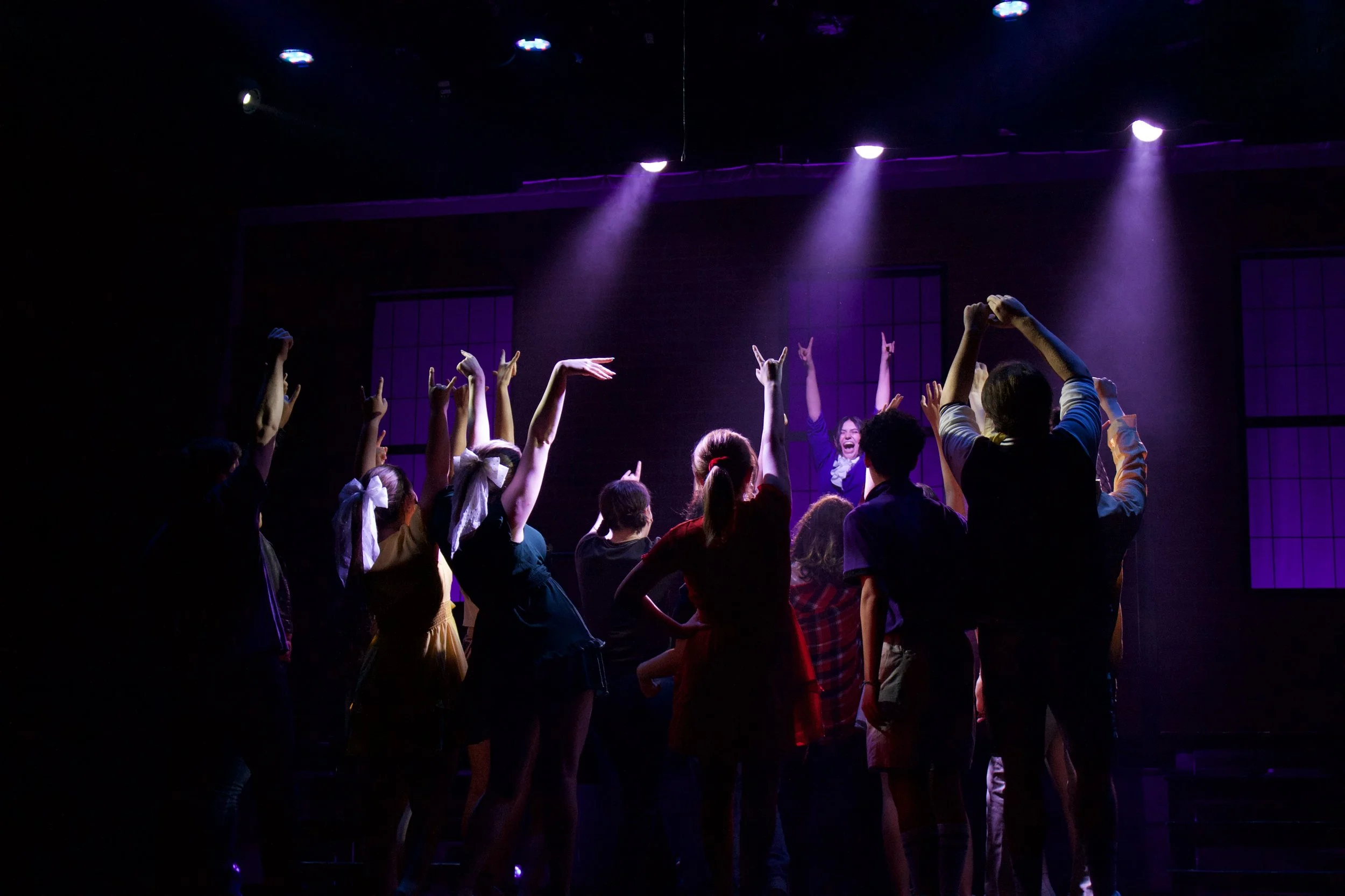 A group of young people on stage participating in a performance, with purple stage lights shining down and some wearing retro or vintage clothing.