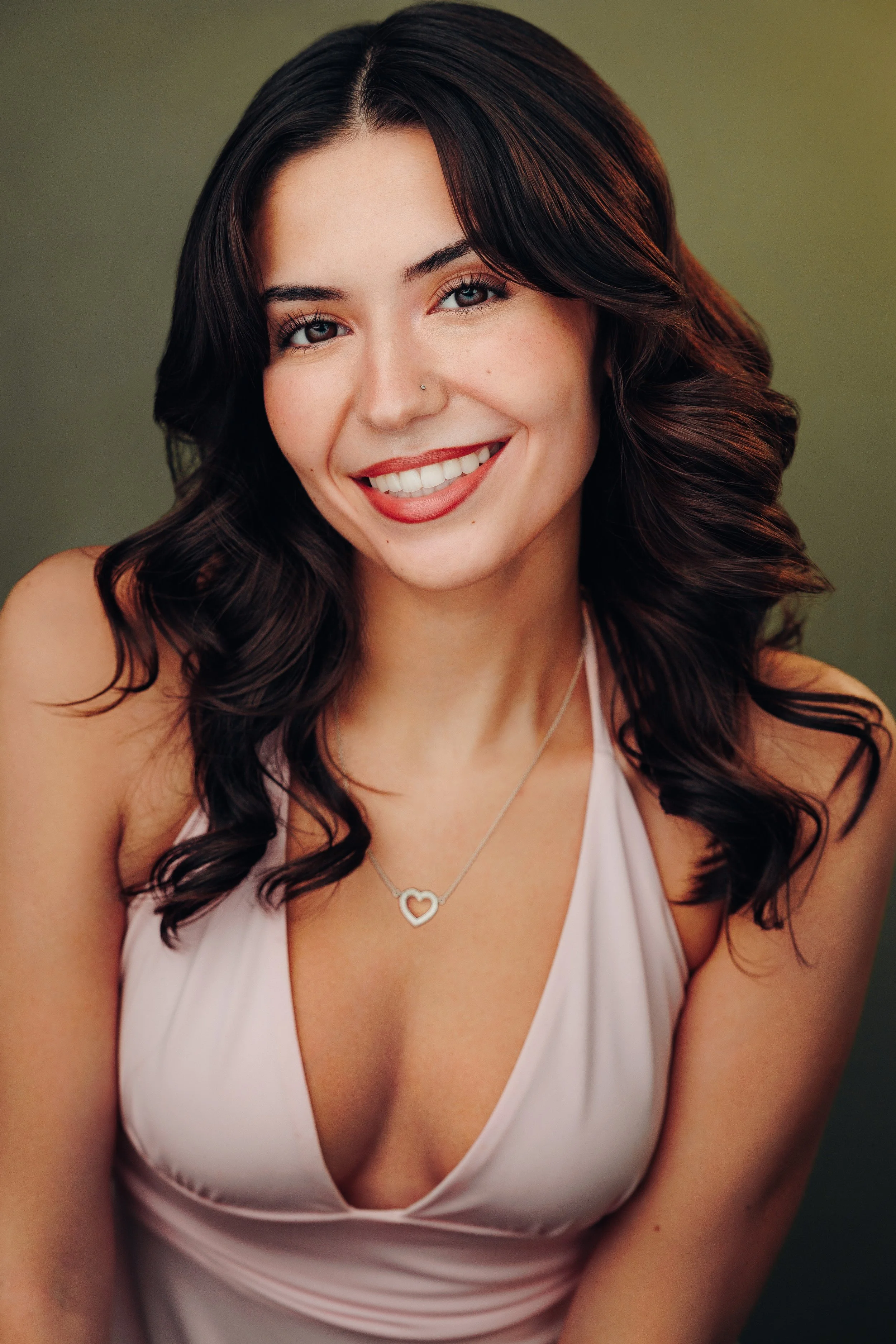 A young woman with long, dark hair and a bright smile, wearing a red textured top and a heart-shaped necklace, standing against a light blue background.