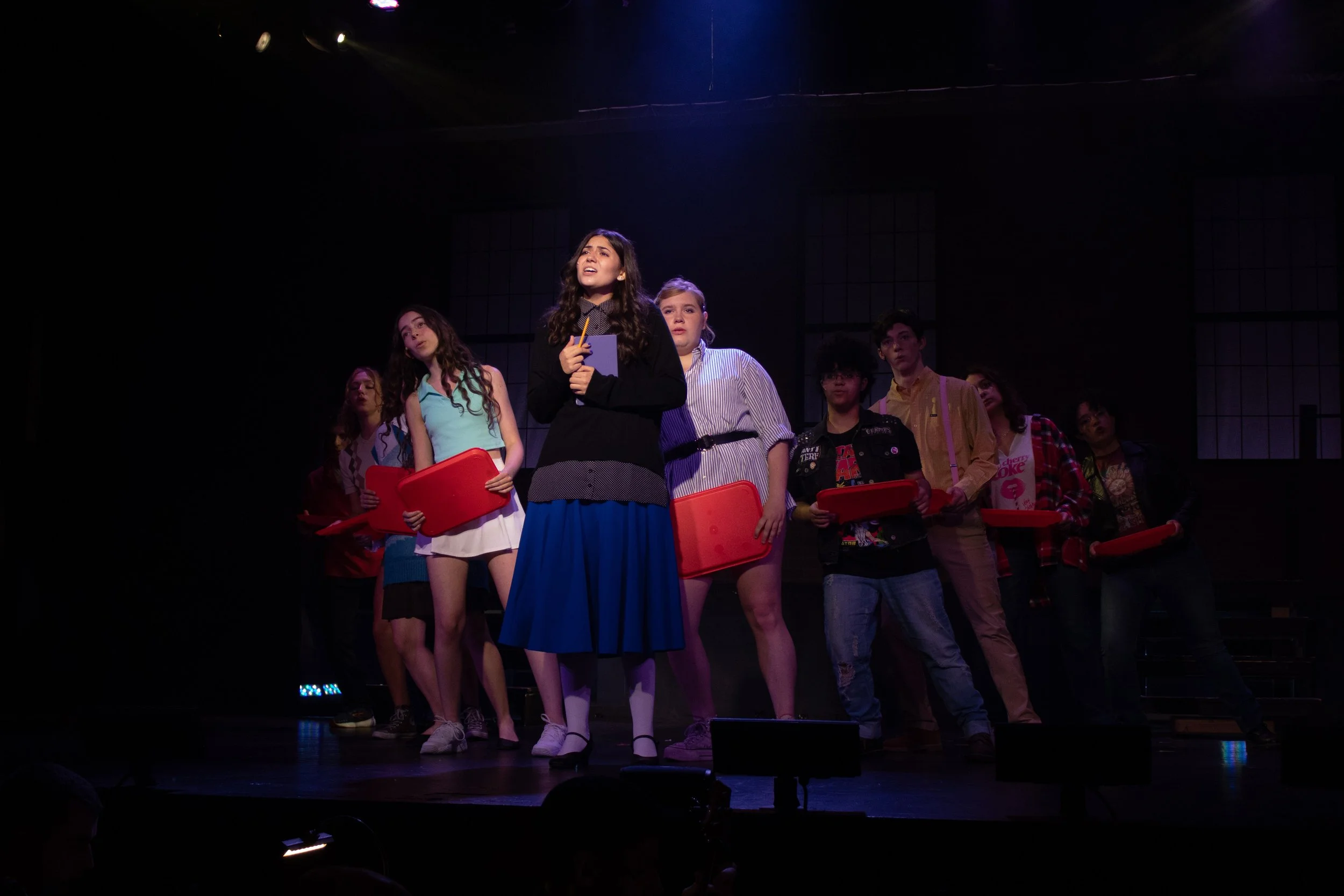 A group of young people standing on a dimly lit stage, some holding red scripts, with one girl in front speaking or singing, possibly a school or community theater performance.