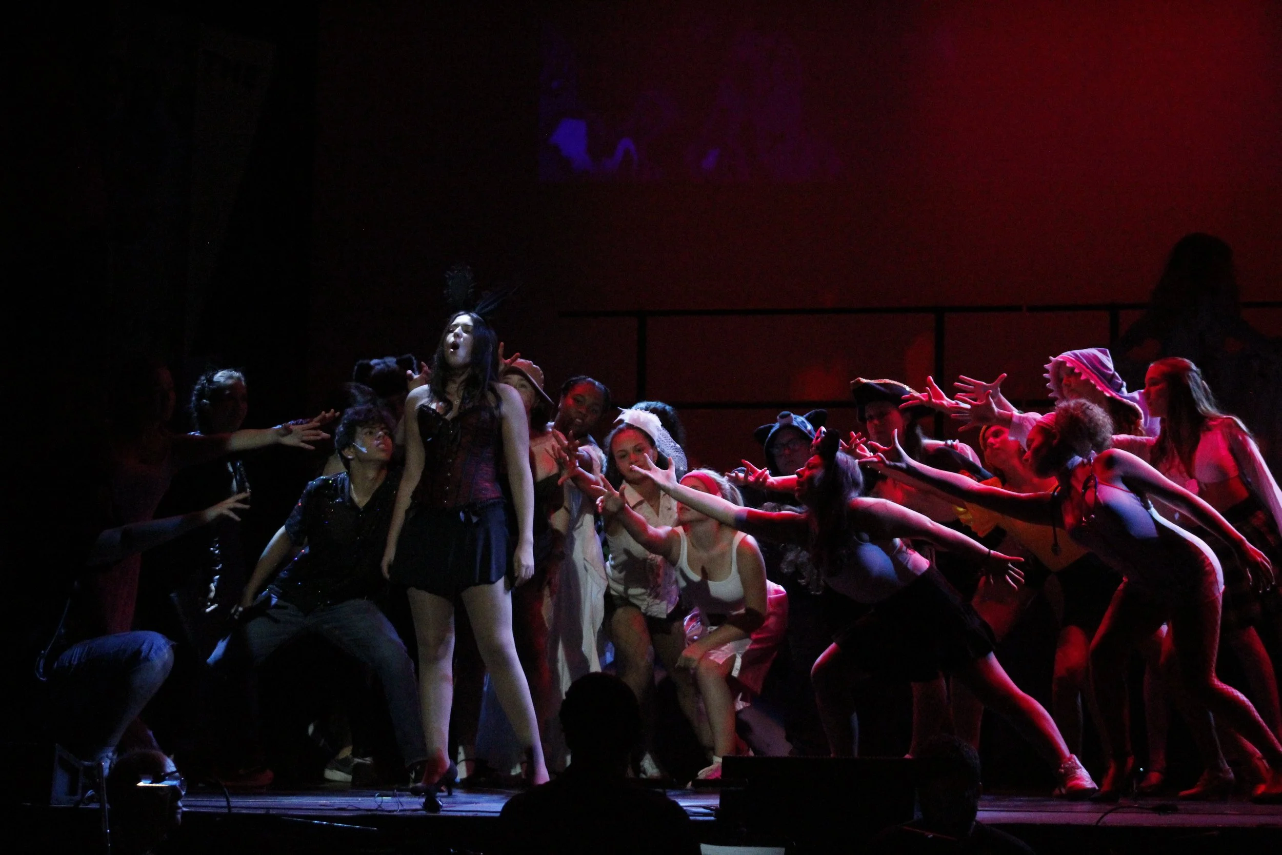 The image shows a theatrical stage performance with a group of young female performers. The lighting is dark with some red hues. The performers are dressed in colorful costumes and are reaching out towards a girl in the center who wears a dark, Victorian-style dress, a headpiece with feathers, and has her mouth open as if singing or speaking. The scene has a dramatic, fantastical atmosphere with the performers' expressive gestures.