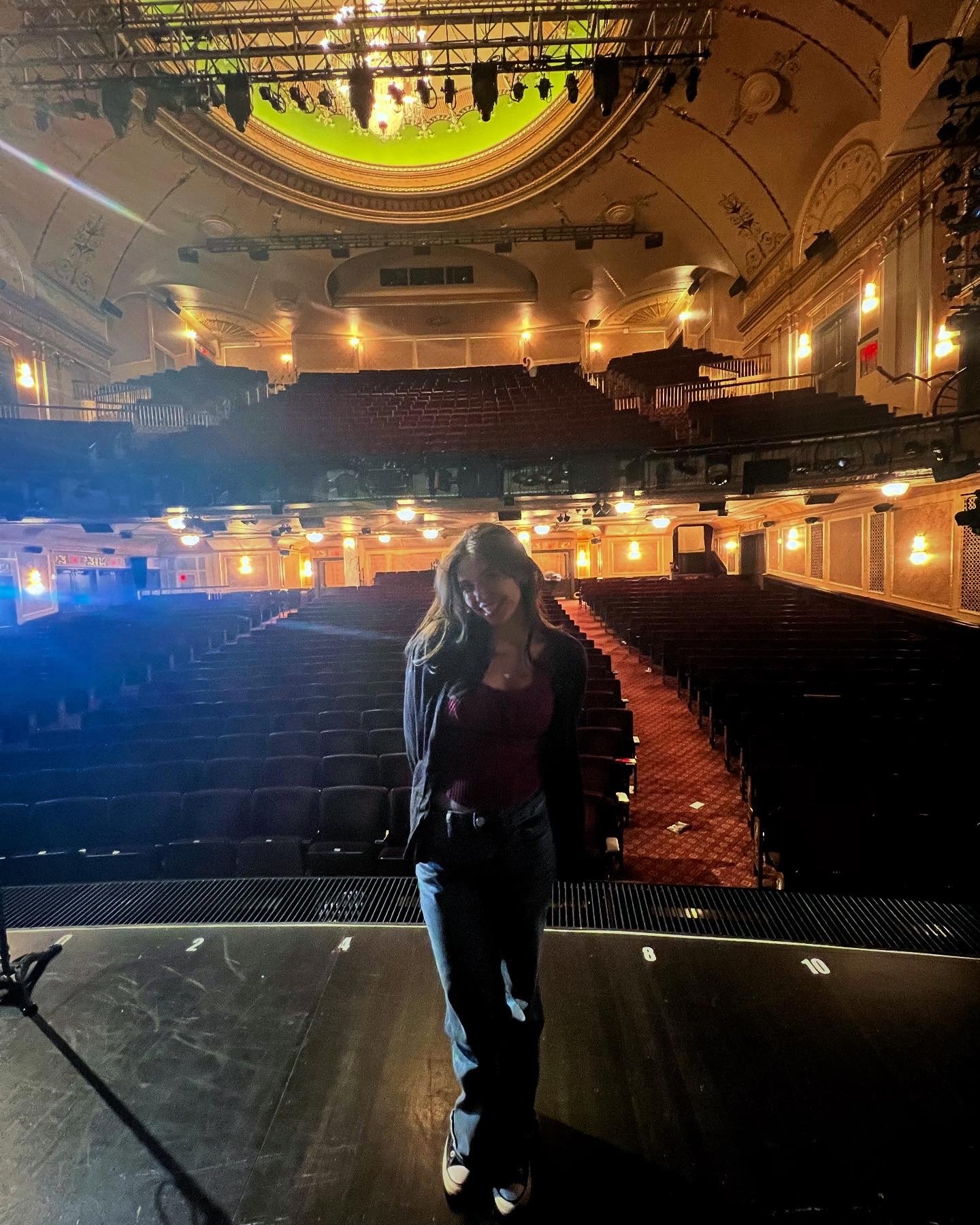 A woman standing on a stage in an ornate theater with empty seats, decorative ceiling, and stage lighting.