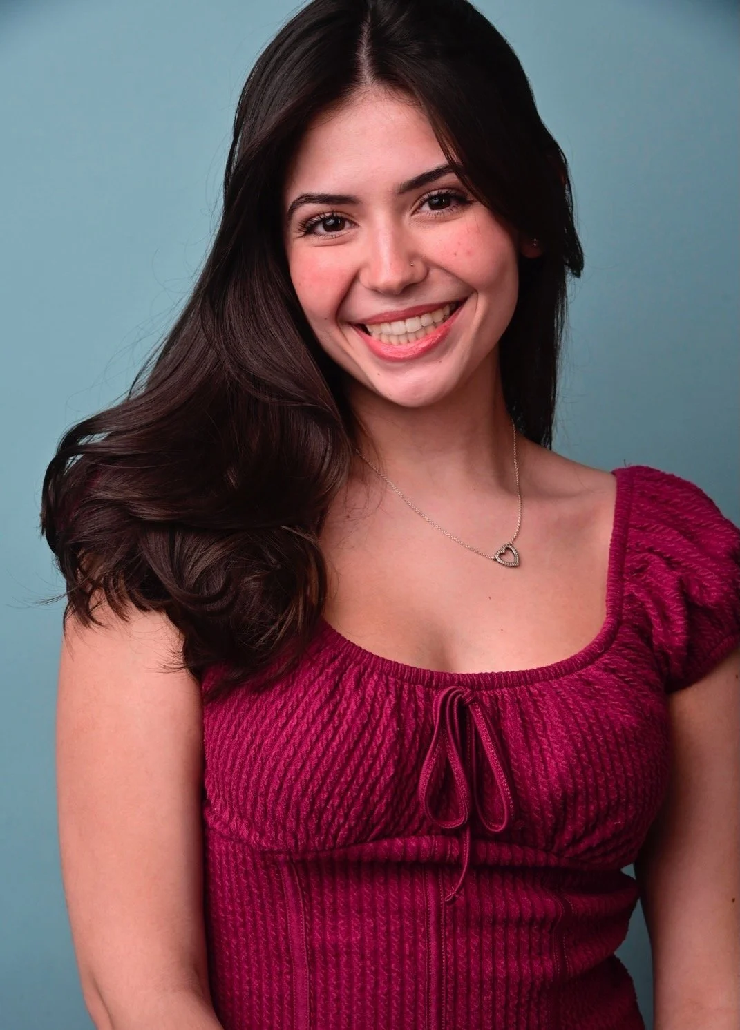 A young woman with long, dark hair and a bright smile, wearing a red textured top and a heart-shaped necklace, standing against a light blue background.