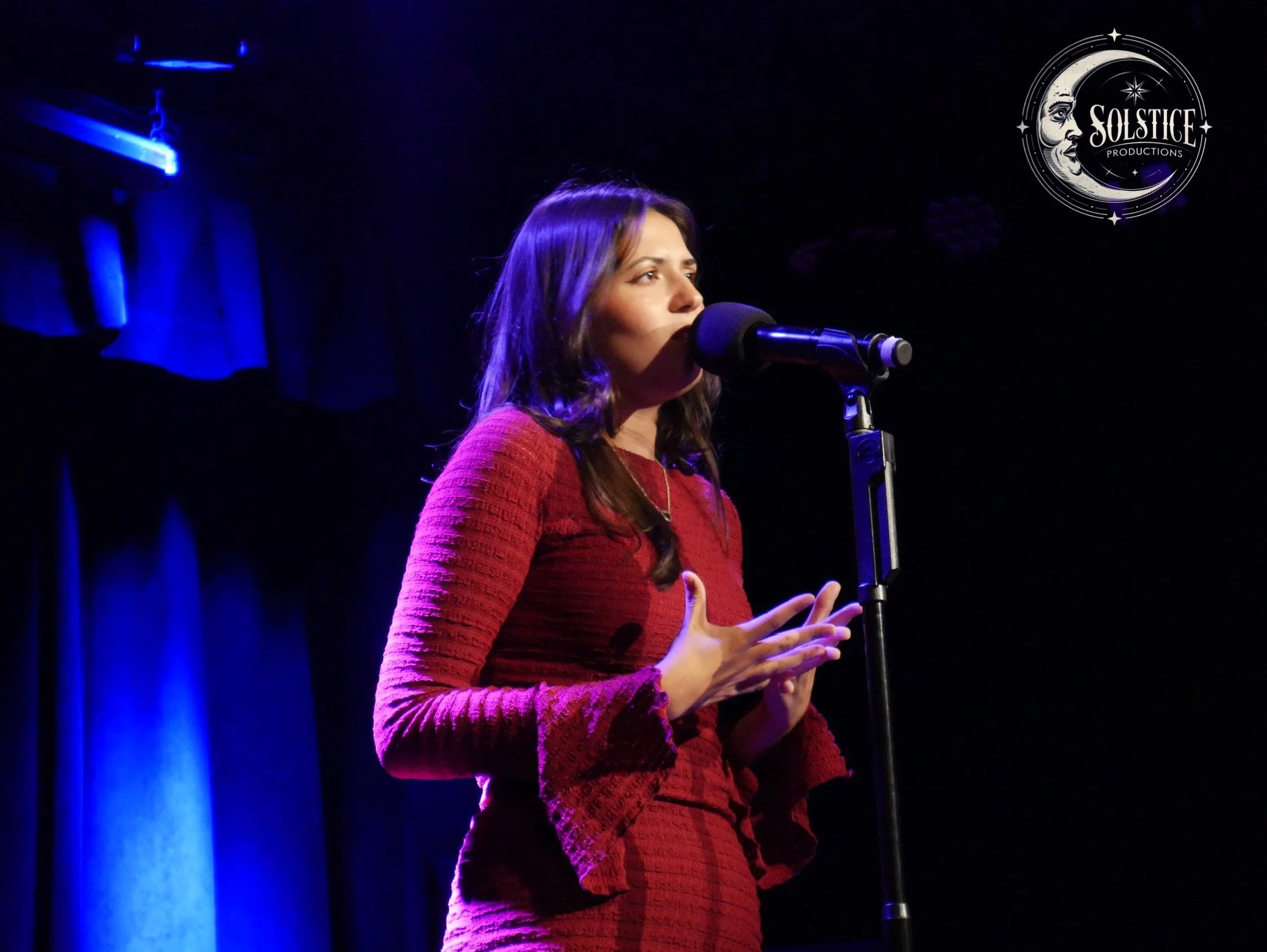A woman in a red textured dress speaking into a microphone on stage with blue lighting and a black curtain background.