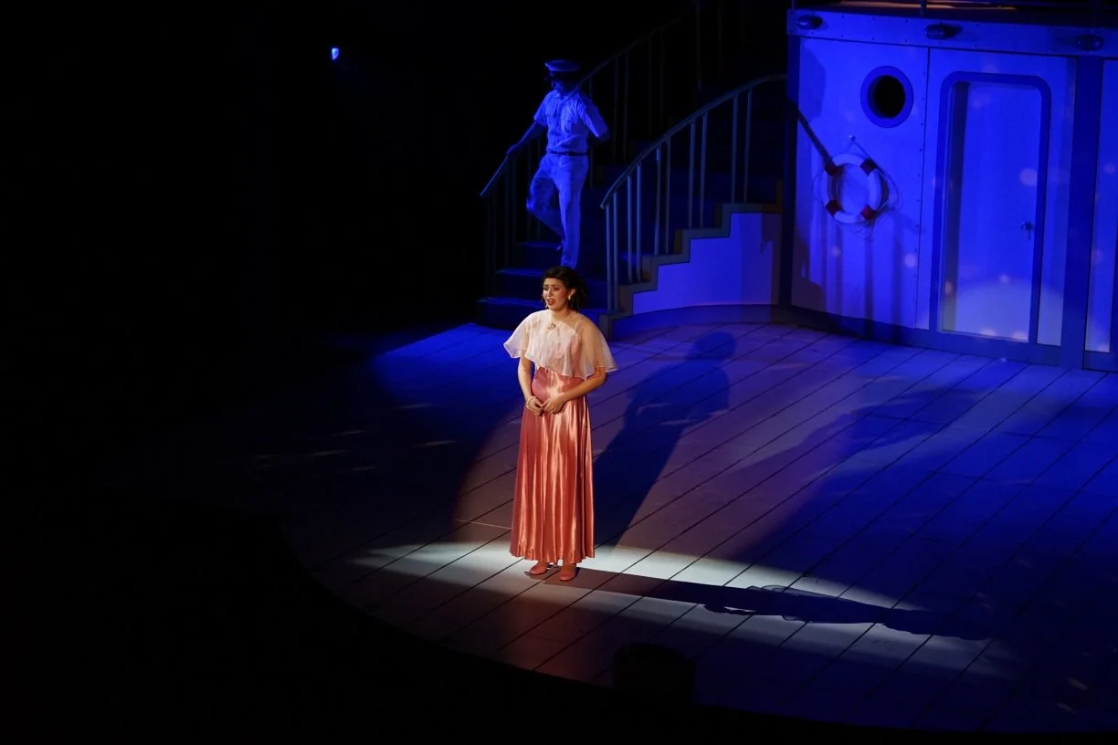 A woman in a peach-colored satin skirt and beige blouse is singing on a stage illuminated by a spotlight. There is a man in a blue uniform standing on stairs behind her, and the stage has nautical-themed decor with a life preserver and blue lighting.