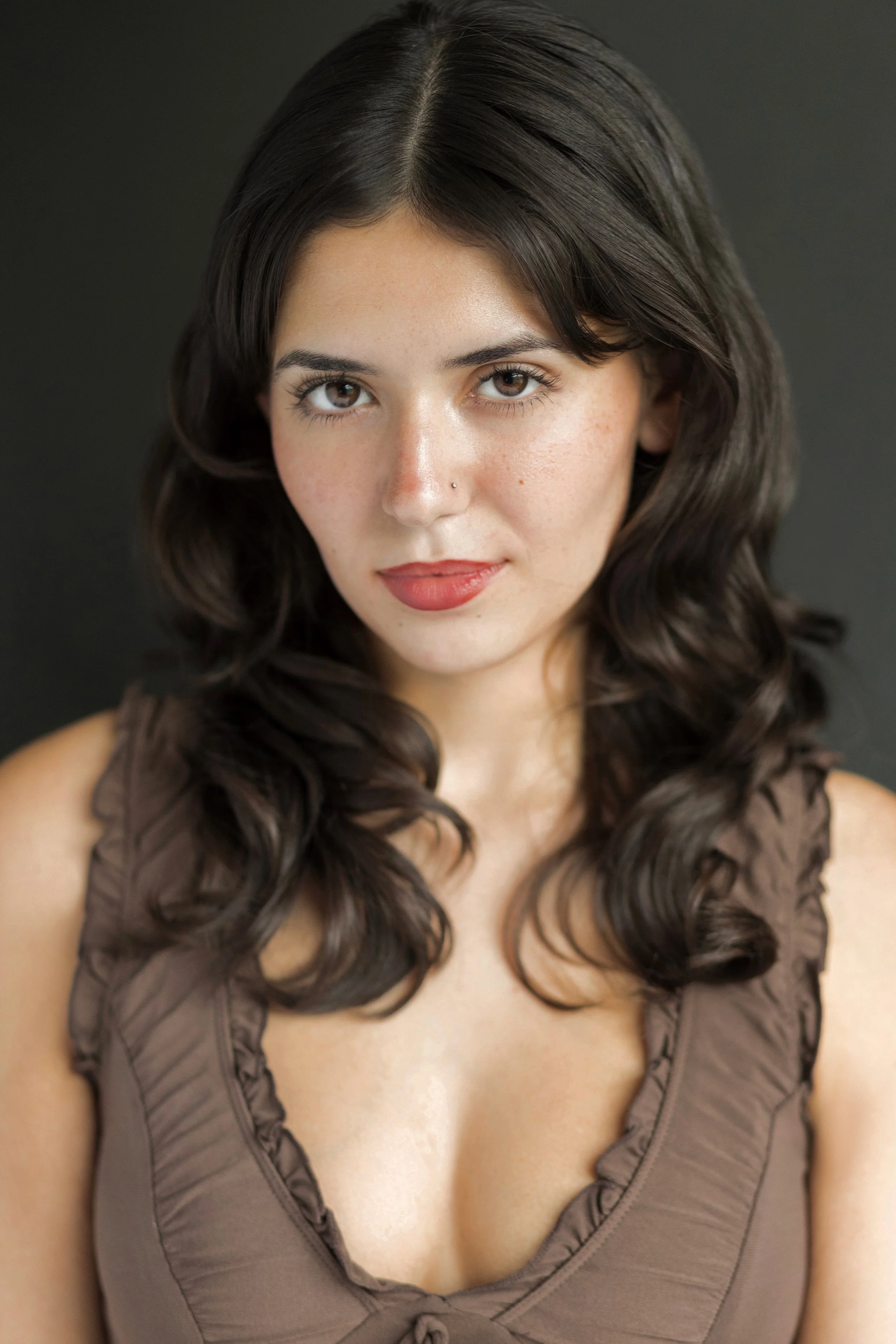 Close-up portrait of a young woman with brown wavy hair, brown eyes, wearing a blue lace top and a blue beaded necklace with a teardrop pendant, standing in front of a red brick wall.