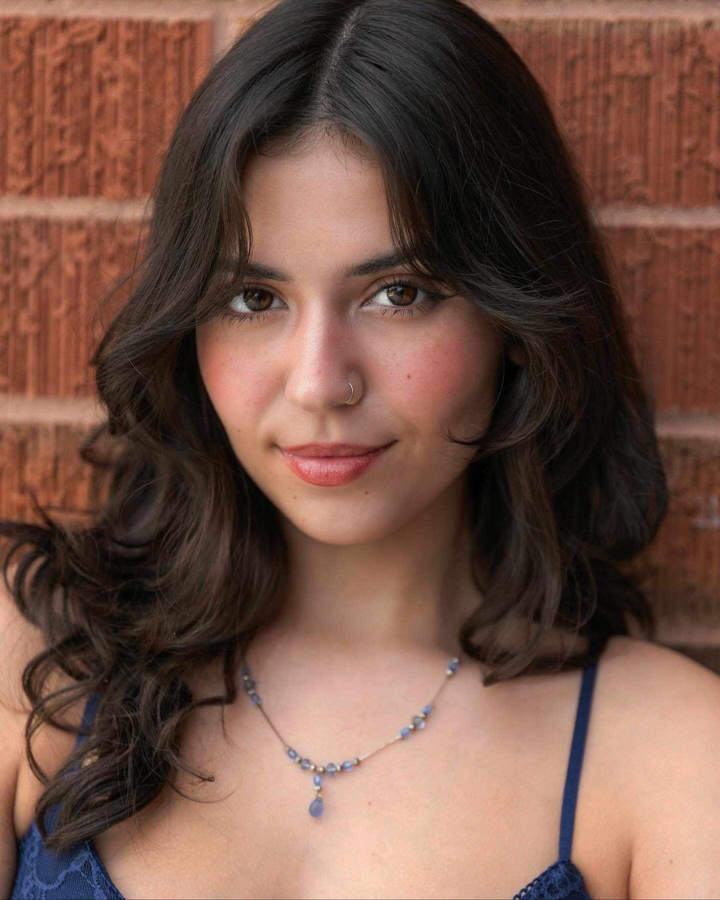 Close-up portrait of a young woman with brown wavy hair, brown eyes, wearing a blue lace top and a blue beaded necklace with a teardrop pendant, standing in front of a red brick wall.