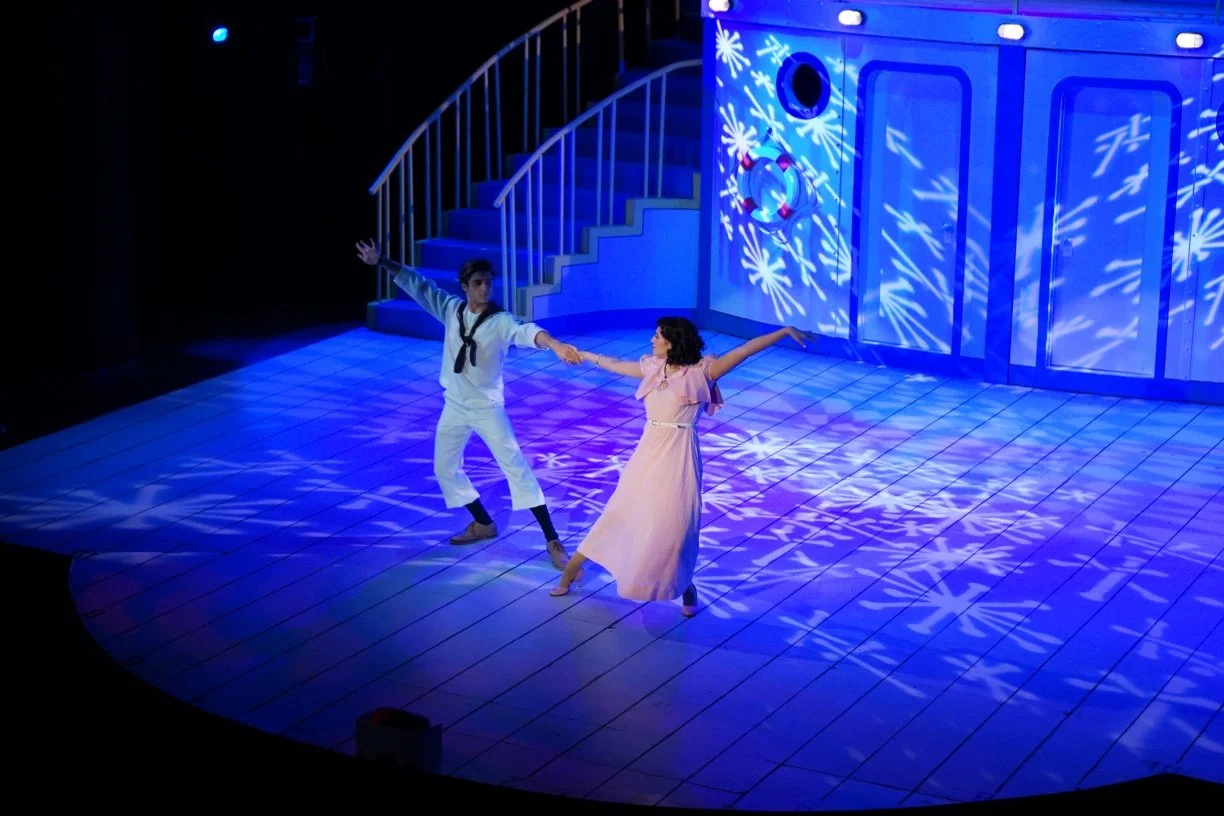 A scene from a stage performance showing a man dressed as a sailor and a woman in a pink dress dancing together on a wooden stage with blue lighting and snowflake patterns projected on the background.