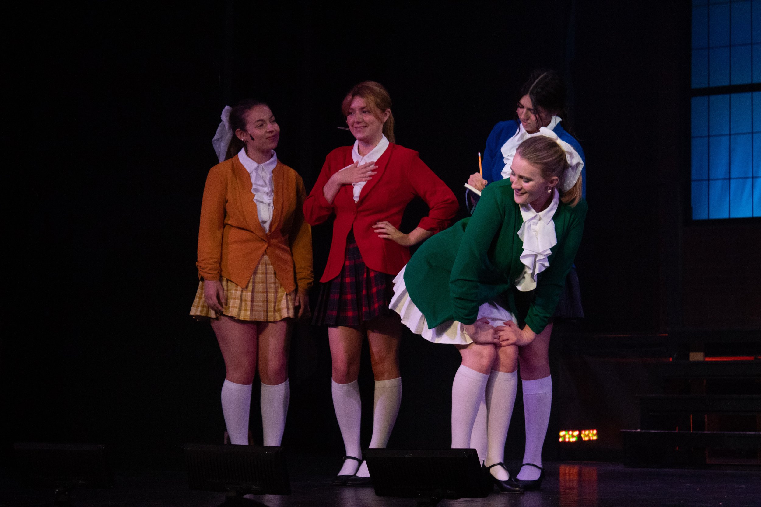Group of four women on stage, dressed in colorful outfits, with one bending down and the others standing, with a dark background and stage lighting.
