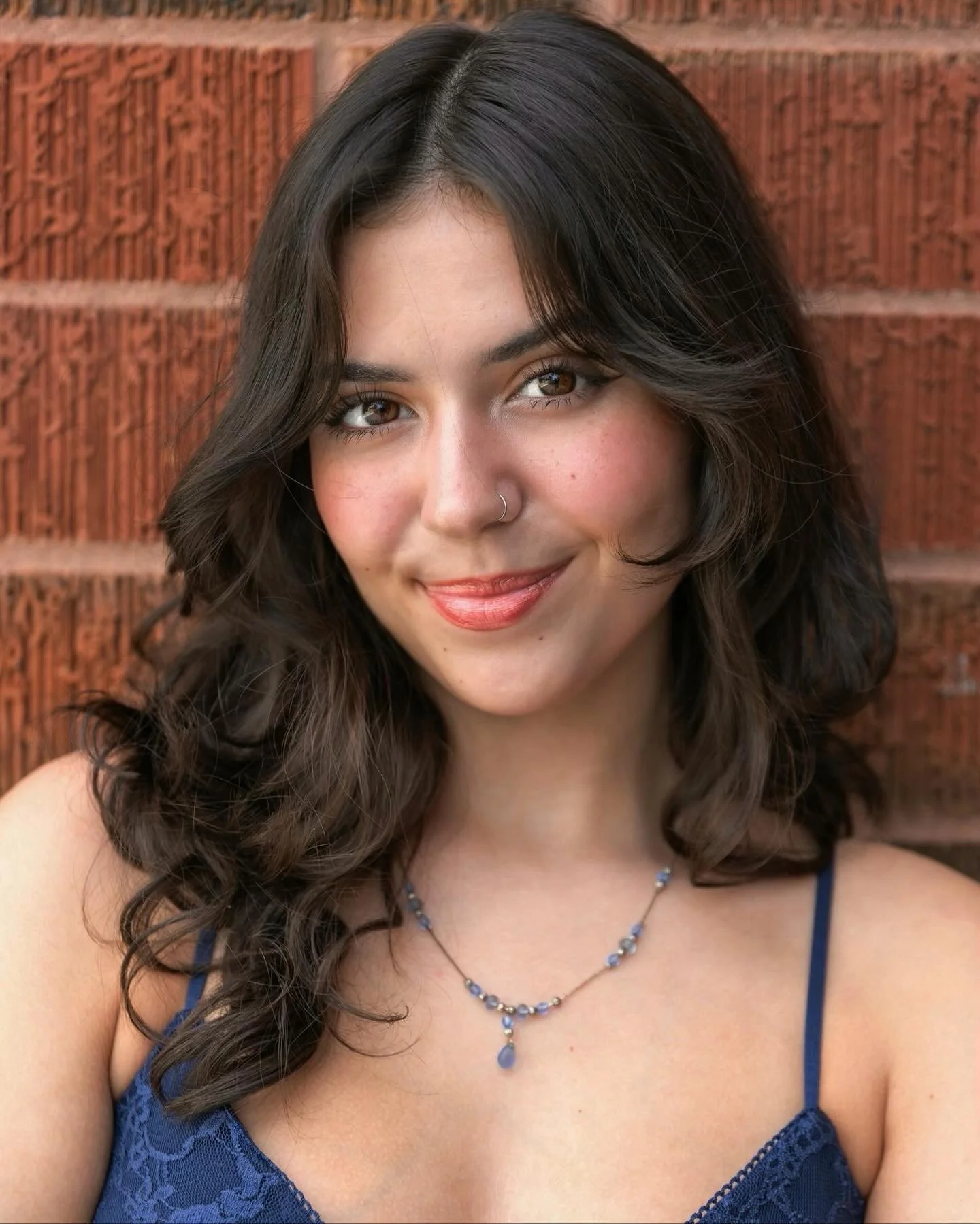 Close-up portrait of a young woman with wavy dark brown hair, brown eyes, wearing a blue lace top and a silver necklace, standing in front of a red brick wall.