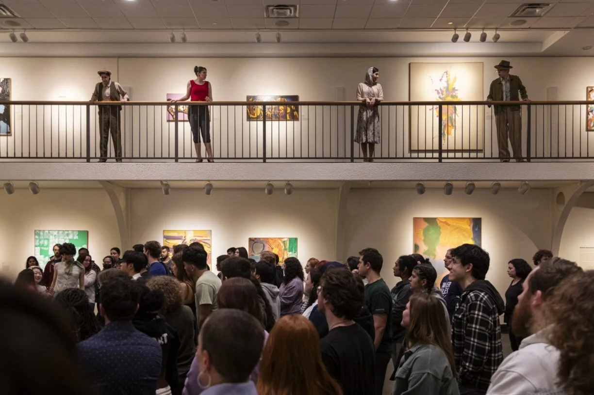 Gallery visitors observing artwork. People standing on a balcony above looking down at crowd in an art exhibition, with colorful paintings on the walls.