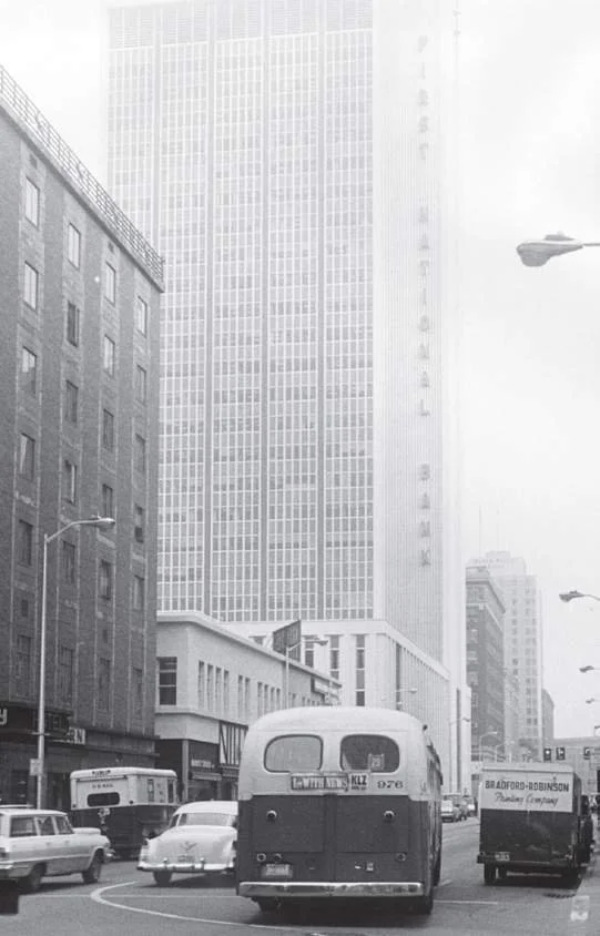 Black and white photo of a busy city street with tall buildings, cars, and trucks, including a vintage bus in the center.
