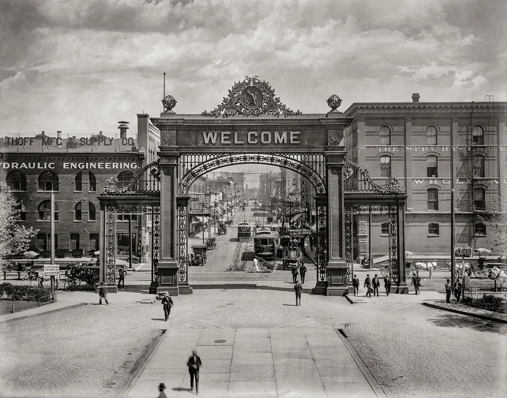 Black and white photograph of a historic city street scene with an ornate gate that says "Welcome," various buildings, streetcars, and pedestrians.