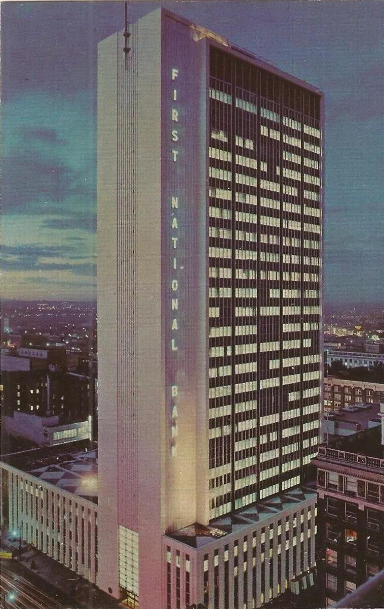 Tall modern high-rise building identified as the First National Bank with large vertical signage, in an urban cityscape at dusk.