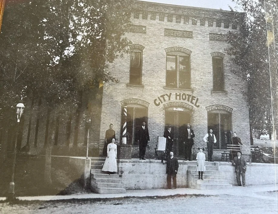 Black and white photo of a vintage brick building labeled 'City Hotel' with nine people standing on the front steps and around it, dressed in early 20th-century attire.