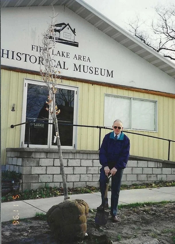 A man wearing sunglasses and a blue jacket holding a shovel, standing in front of the Five Lake Area Historical Museum. There is a small, leafless tree planted in a burlap-wrapped root ball nearby.
